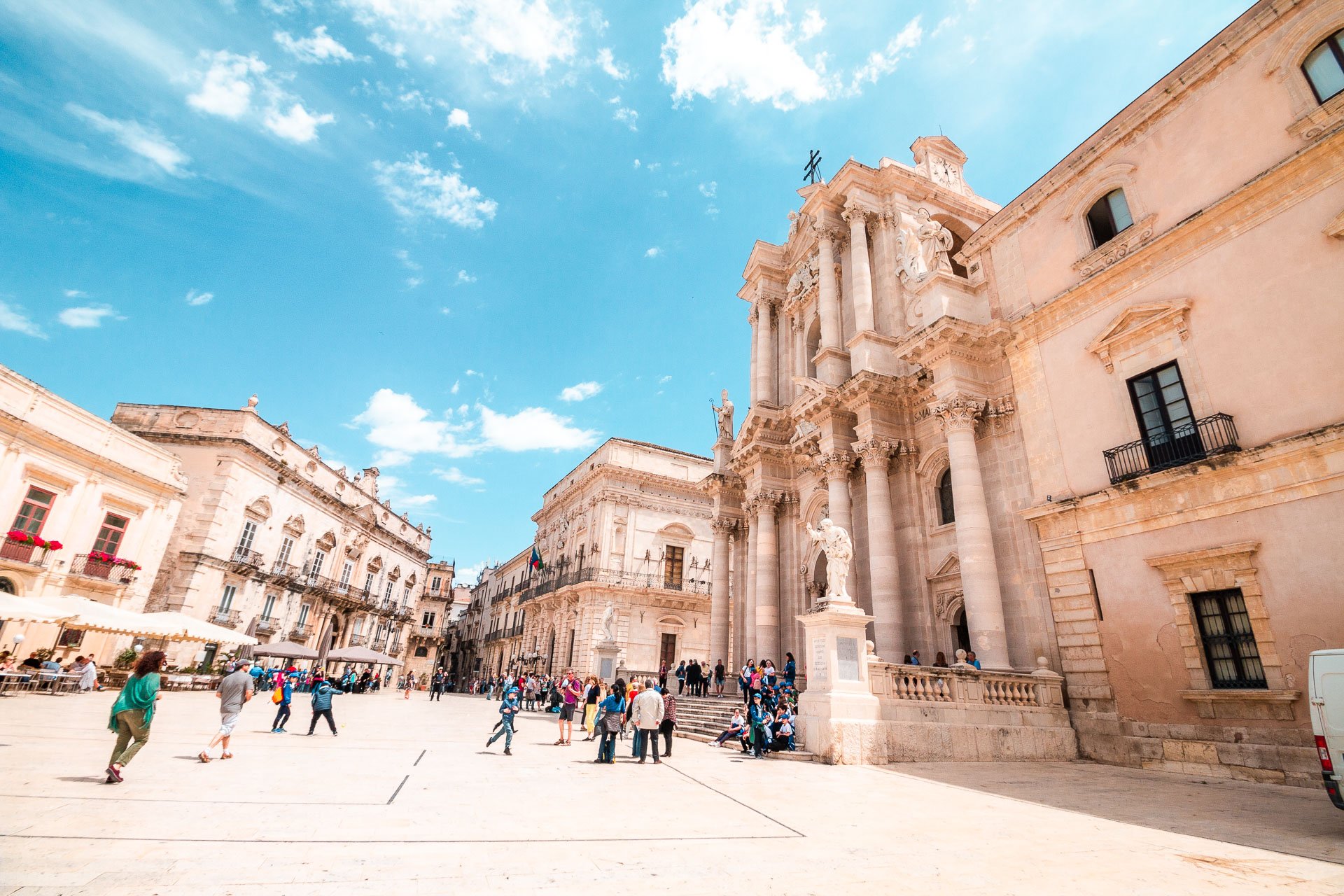 This image shows Piazza Duomo in Siracusa, Sicily, on a sunny day. There are people walking around the square. 