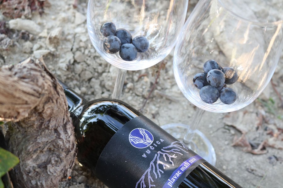 This image shows a bottle of Vucica wine and two empty wine glasses with black grapes in them. 