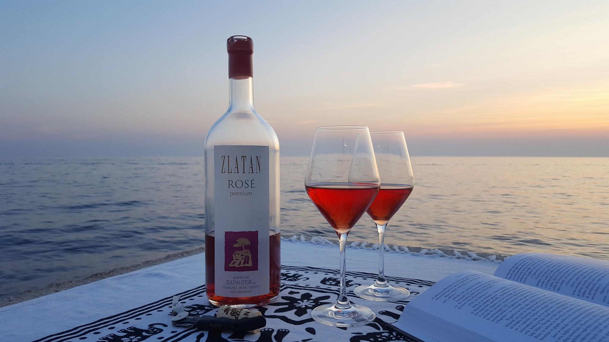 This image shows a half-empty bottle of rose Zlatan Otok wine, two glasses filled with rose wine and an open book with the sea and the sunset in the background. 