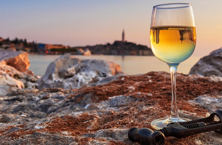 This image shows a glass of white wine on a rock with the town of Rovinj in the background. This is the featured image of our guide to the best wineries in Istria, Croatia.
