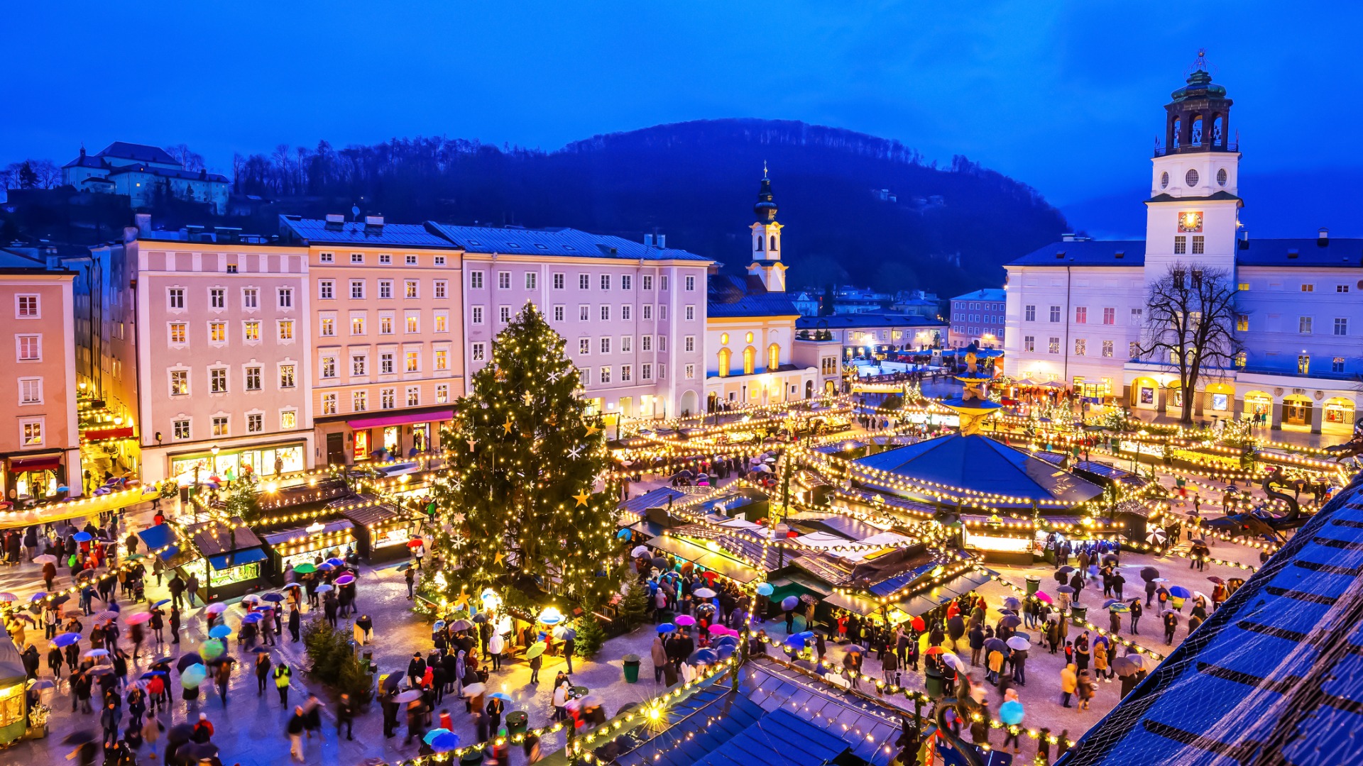 This image shows a panoramic view of Salzburg Christmas Market at dusk. Surrounded by beautiful buildings, the square is filled with stalls and festive lights. There is a Christmas tree in the middle of the square and many people. 