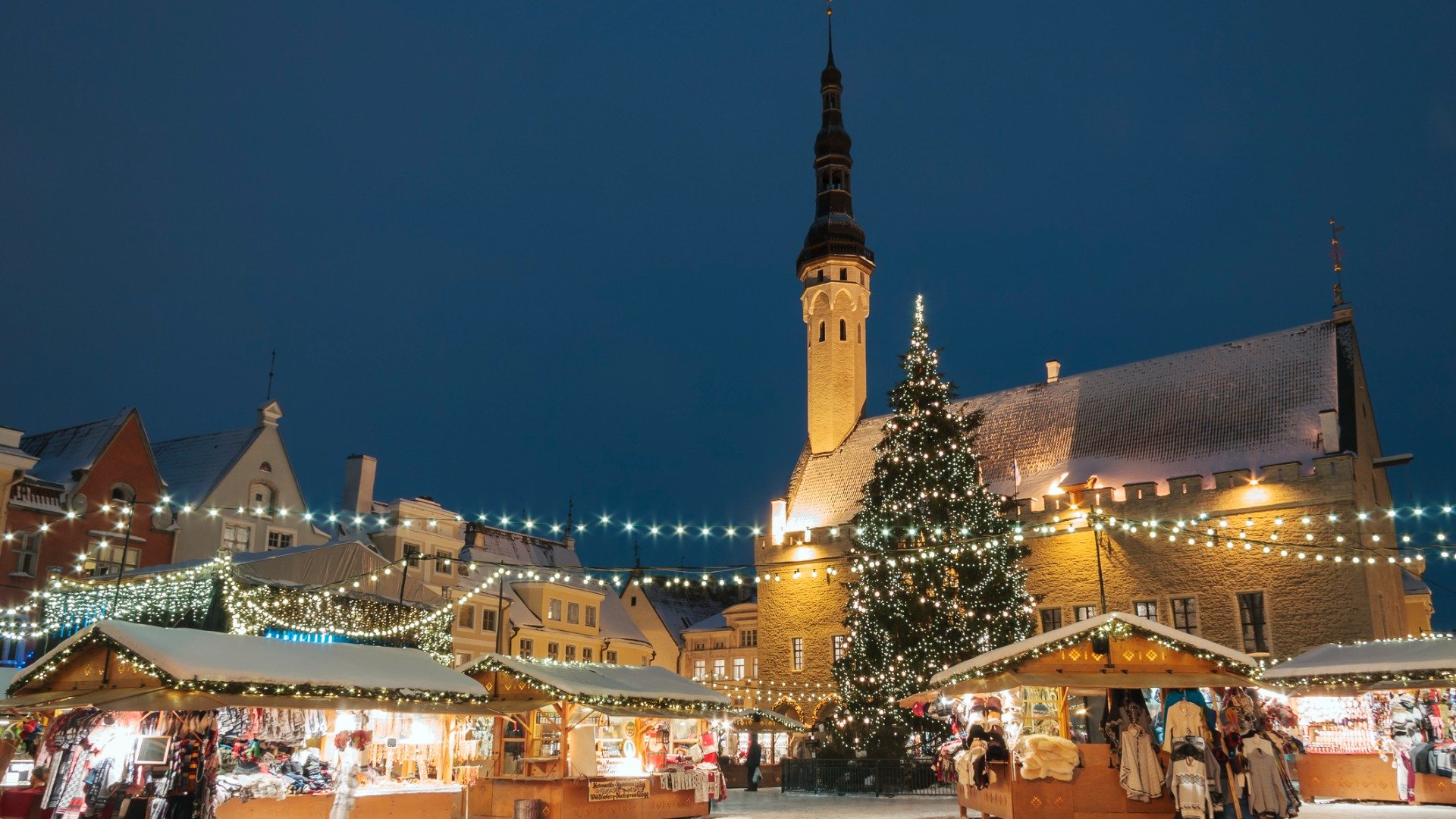 This image shows the Christmas market in Tallinn. There are Christmas lights and people stop by stalls selling souvenirs. 