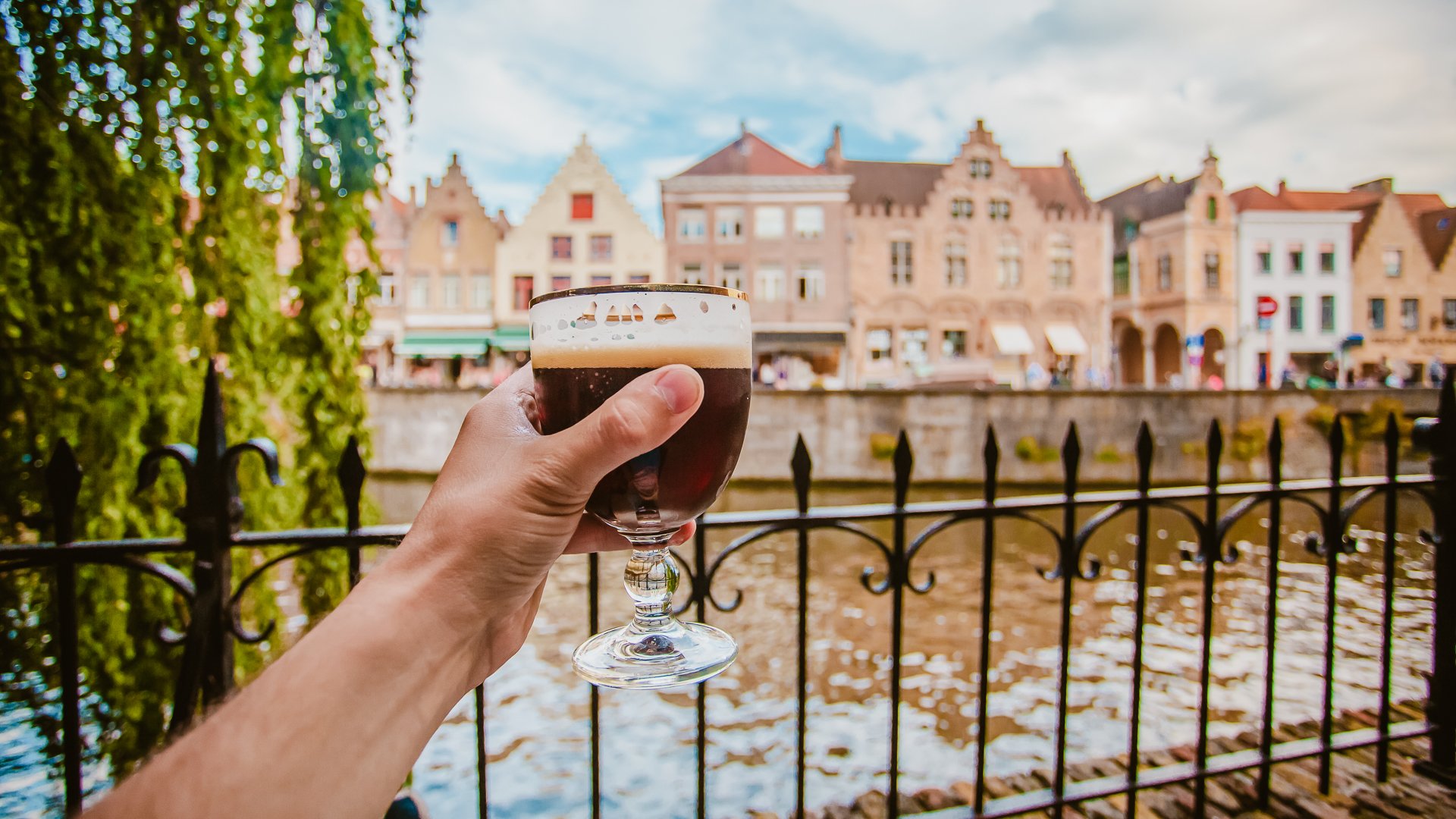 A hand holds a full glass of dark Belgian beer near a canal in Bruges, Belgium with old buildings in the background.