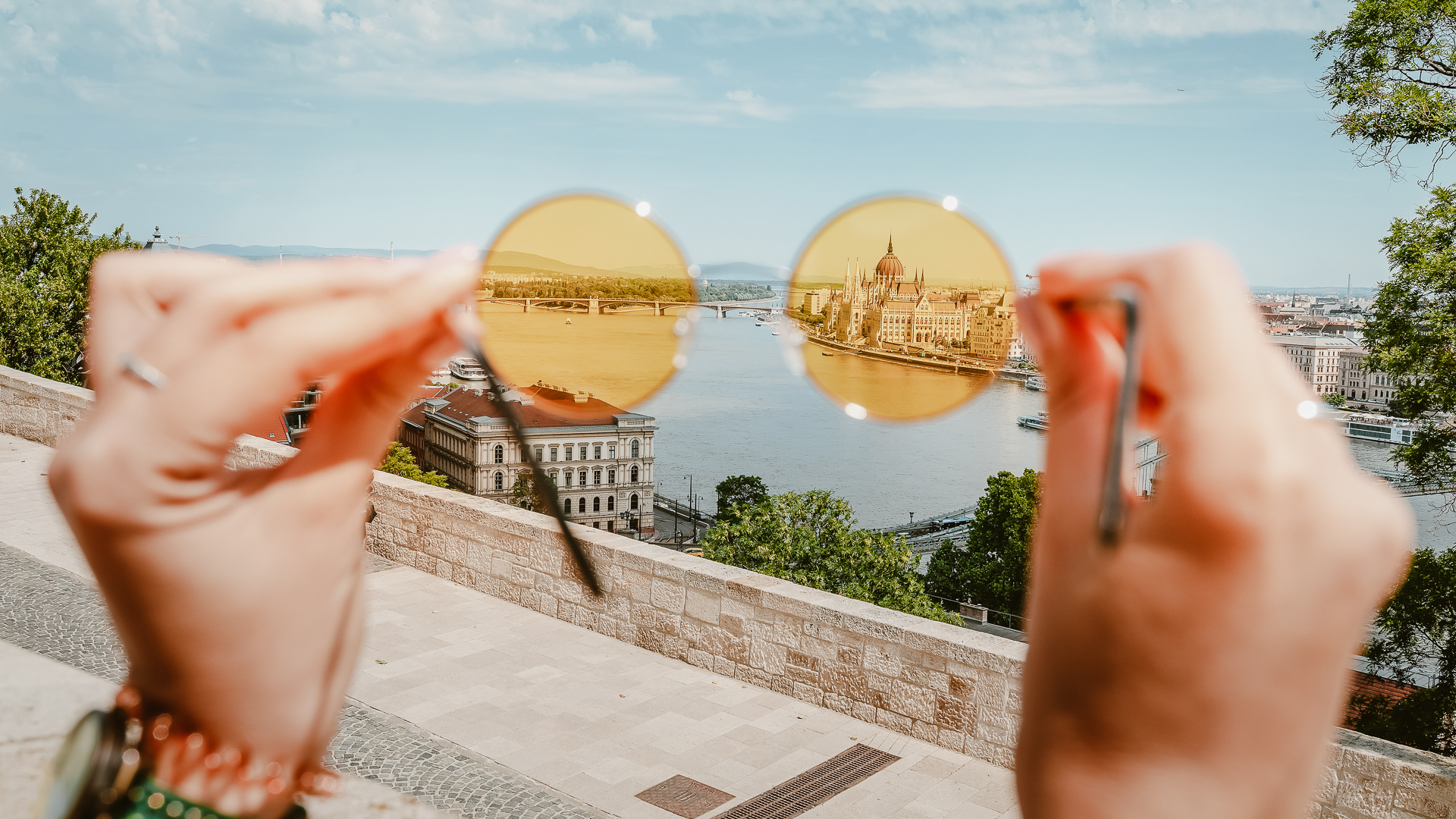 Two hands with funky bracelets hold yellow round hippie glasses out of focus in foreground - in the background, in focus, we see the parliament building in Budapest, Hungary.