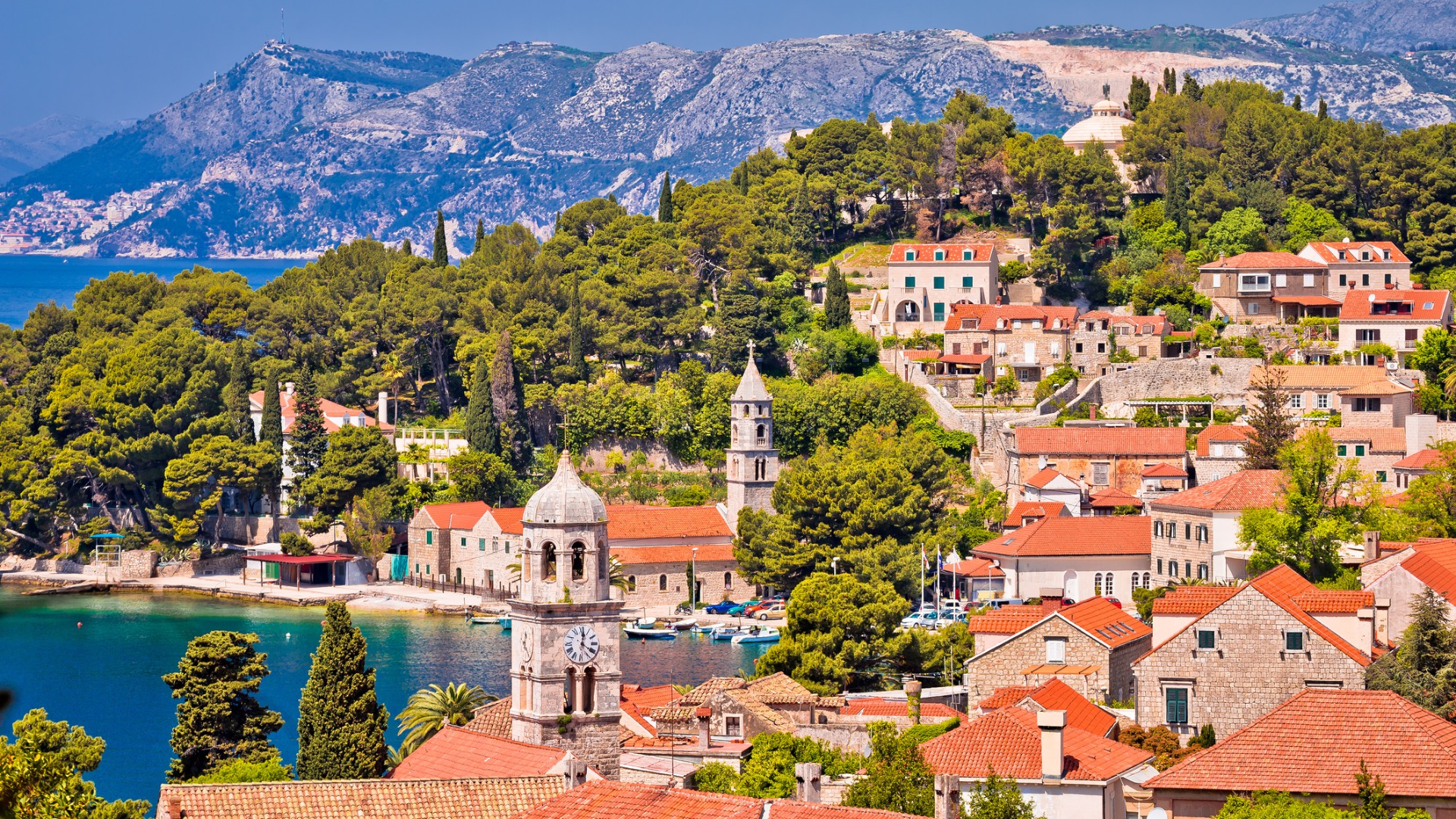 Esta é uma visão panorâmica de Cavtat.  Há casas de pedra com telhados vermelhos construídas entre árvores.  Ao fundo, há uma belíssima praia. 