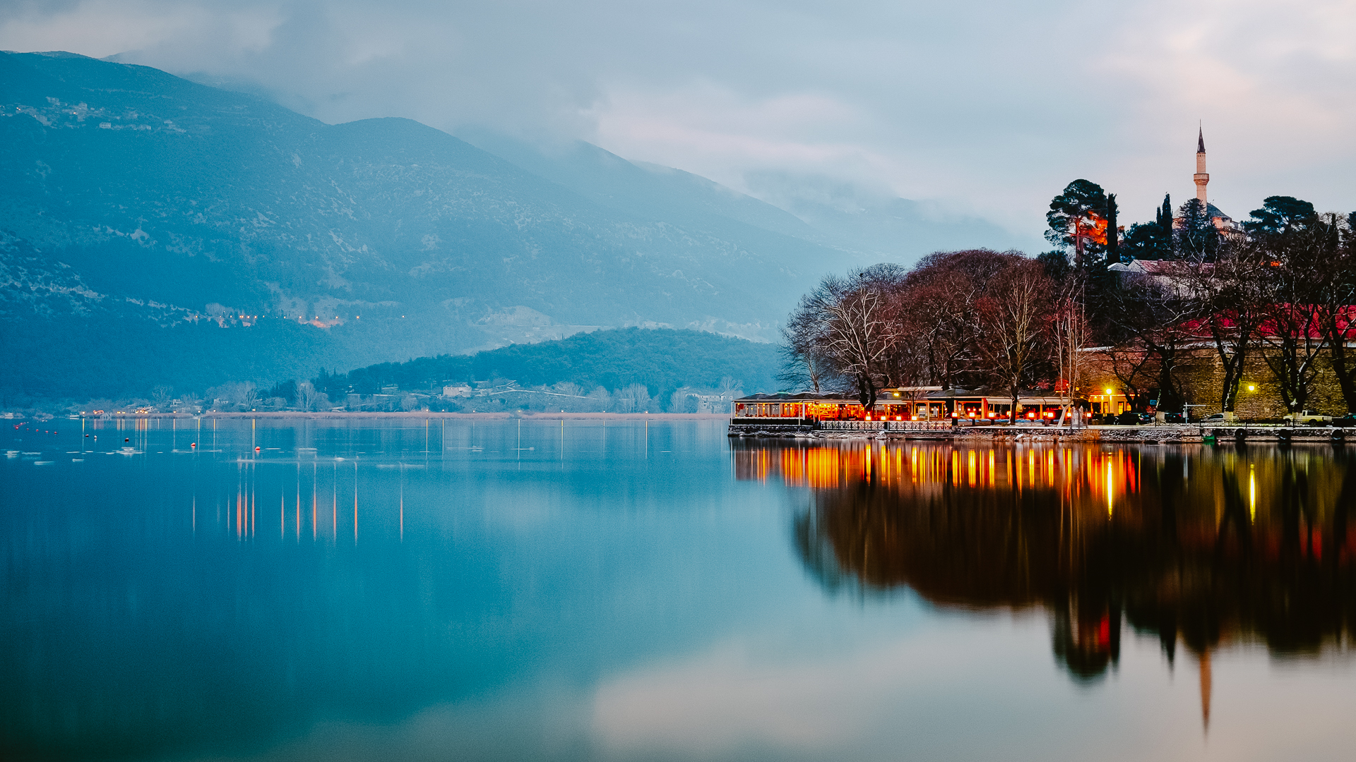 Pamvotida Lake in the evening hours with mountains reflecting in the water in the background and to the right a well-lit peninsula with buildings and the Fethiye Mosque standing out.