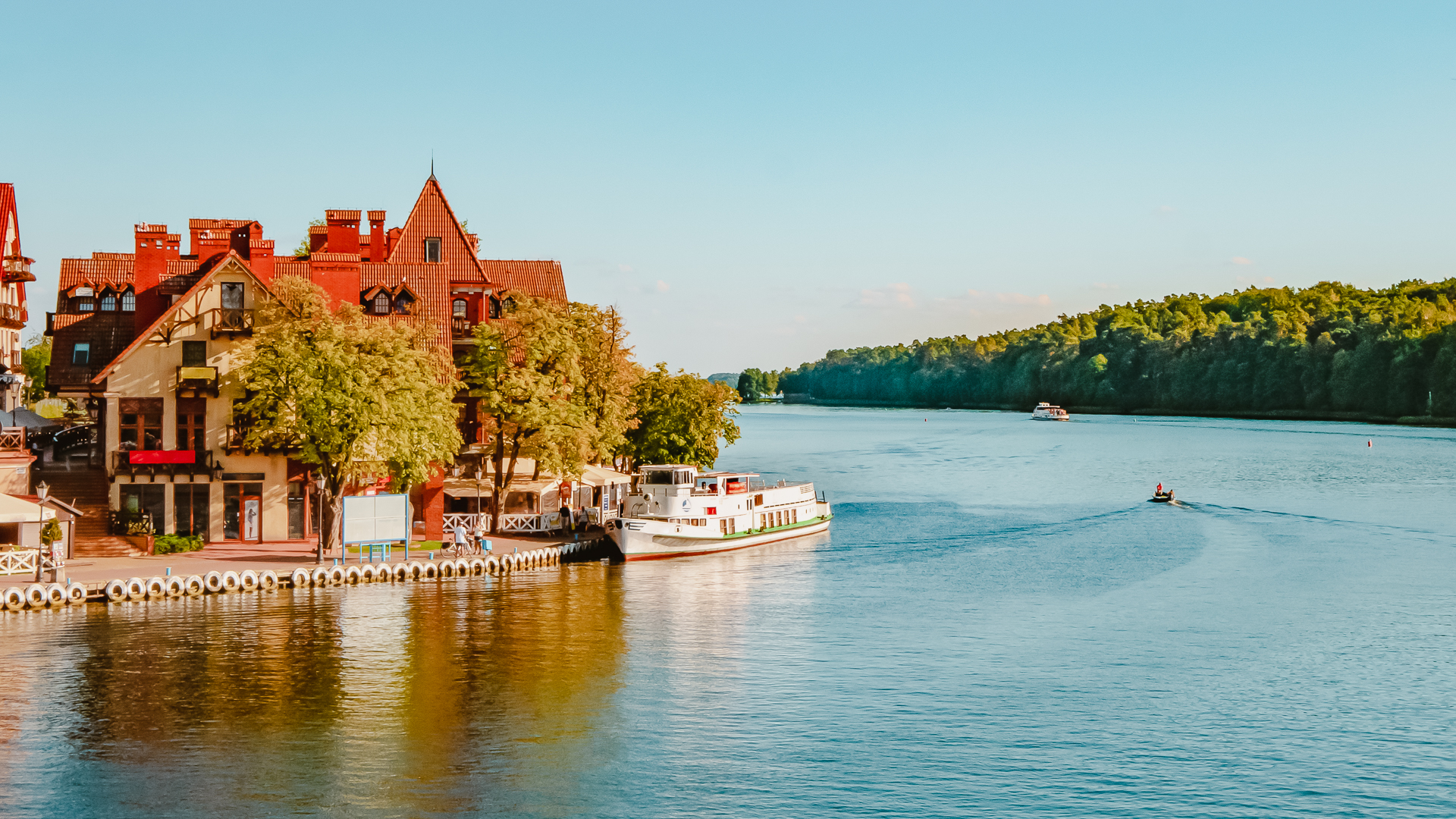 View of a lake with houses on the left side, water in the middle with a small boat passing by and a lush forest to the right in Mikolajki, Poland.