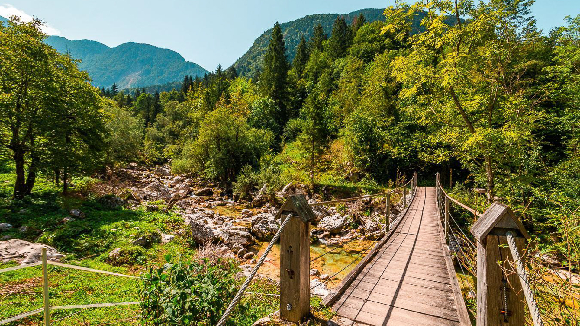 View of a national park in Soca Valley in Slovenia with a wooden suspension bridge on the right and mountains in the background. The scenery is fresh green and lush.