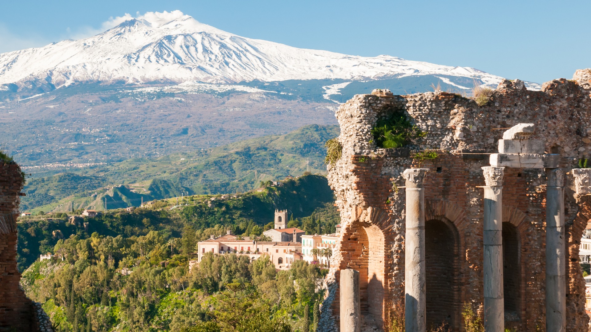 Această imagine prezintă câteva coloane antice din Taormina, cu un munte Etna acoperit de zăpadă în fundal.