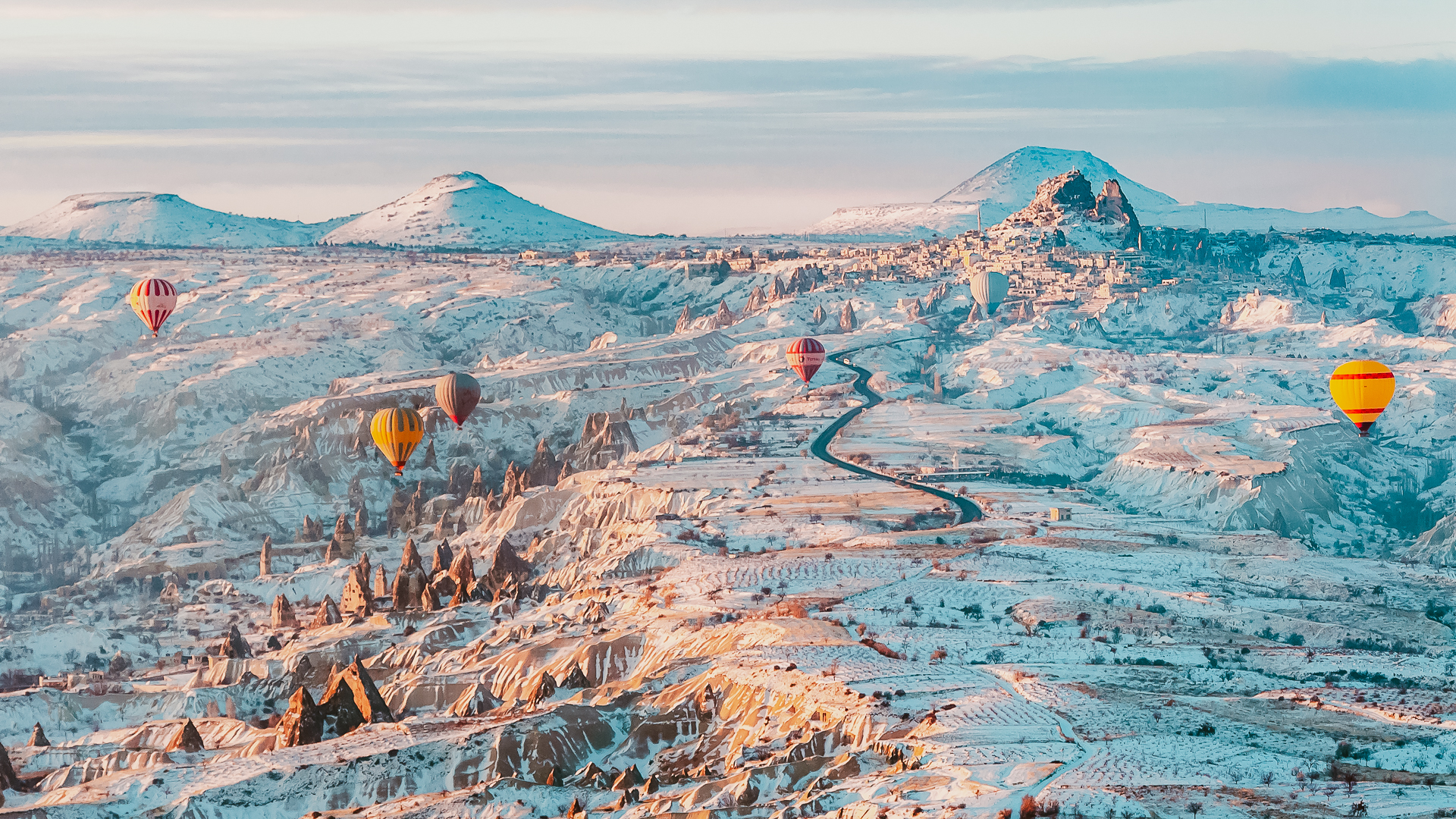 Several warm-colored hot air balloons flying over the snow-covered landscape in Cappadocia, Turkey in golden light conditions.