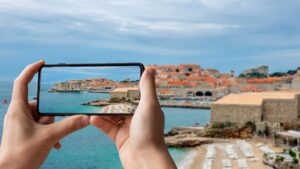 This is a panoramic view of Banje Beach with its turquoise waters and golden sand. In the background, the walled Old Town of Dubrovnik with the iconic red rooftops spreads in all its glory. In the foreground, two hands holding a phone, taking a picture of the landscape.