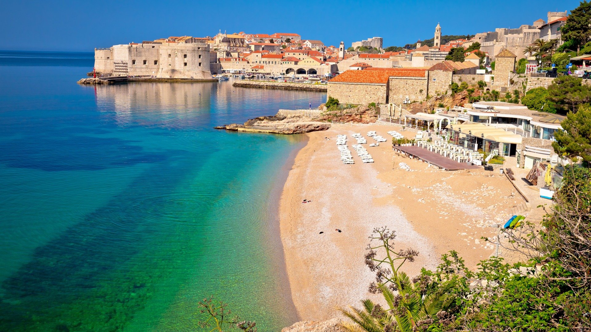 This is a panoramic view of Banje Beach with its turquoise waters and golden sand. In the background, the walled Old Town of Dubrovnik with the iconic red rooftops spreads in all its glory. 