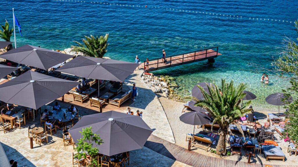 This image shows Cava Beach in Dubrovnik. There are several umbrellas, sun lounges and tables next to the turquoise waters.