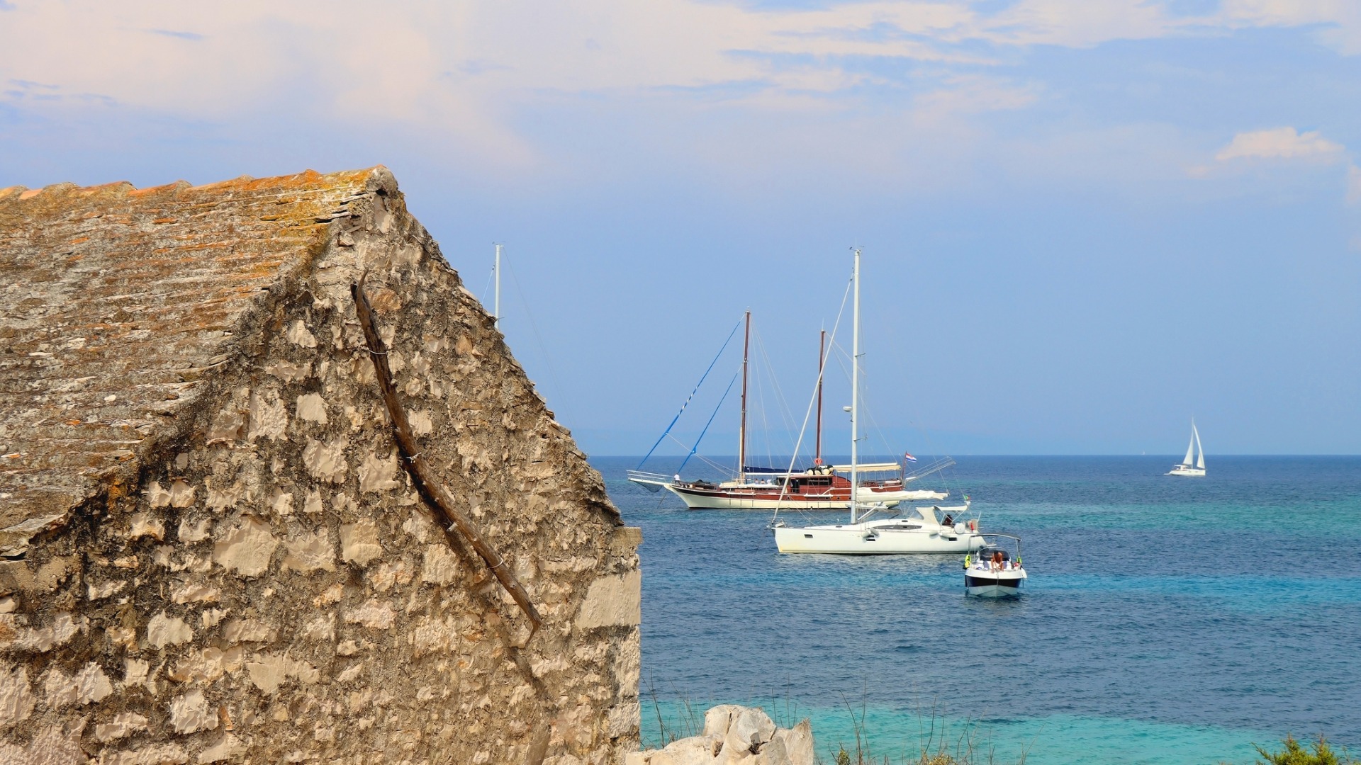 This image shows an old stone hut with the sea and some sailing boats in the background.