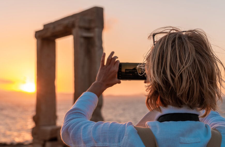 This image shows a woman taking a photo of Portara at sunset with her mobile phone.