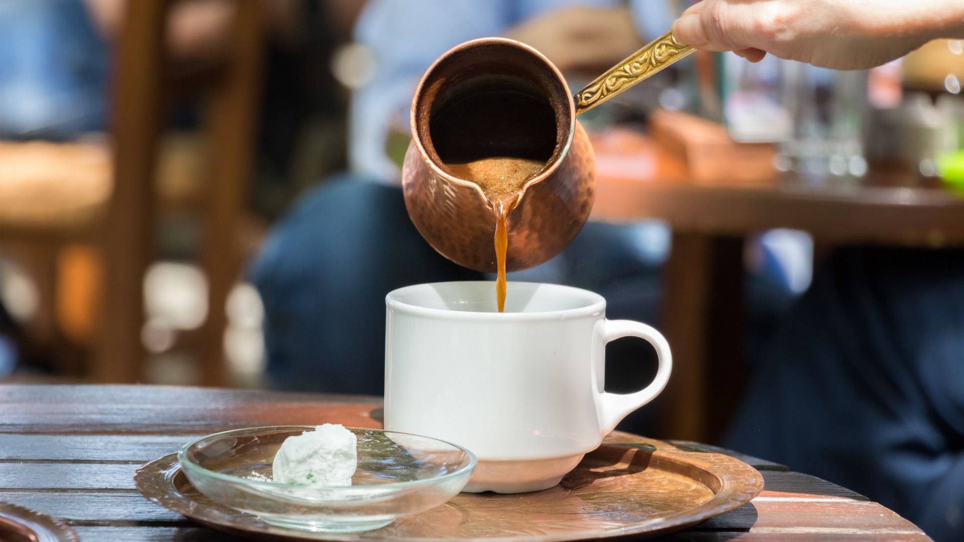 This is a close-up of a woman pouring Greek coffee in a white cup. 
