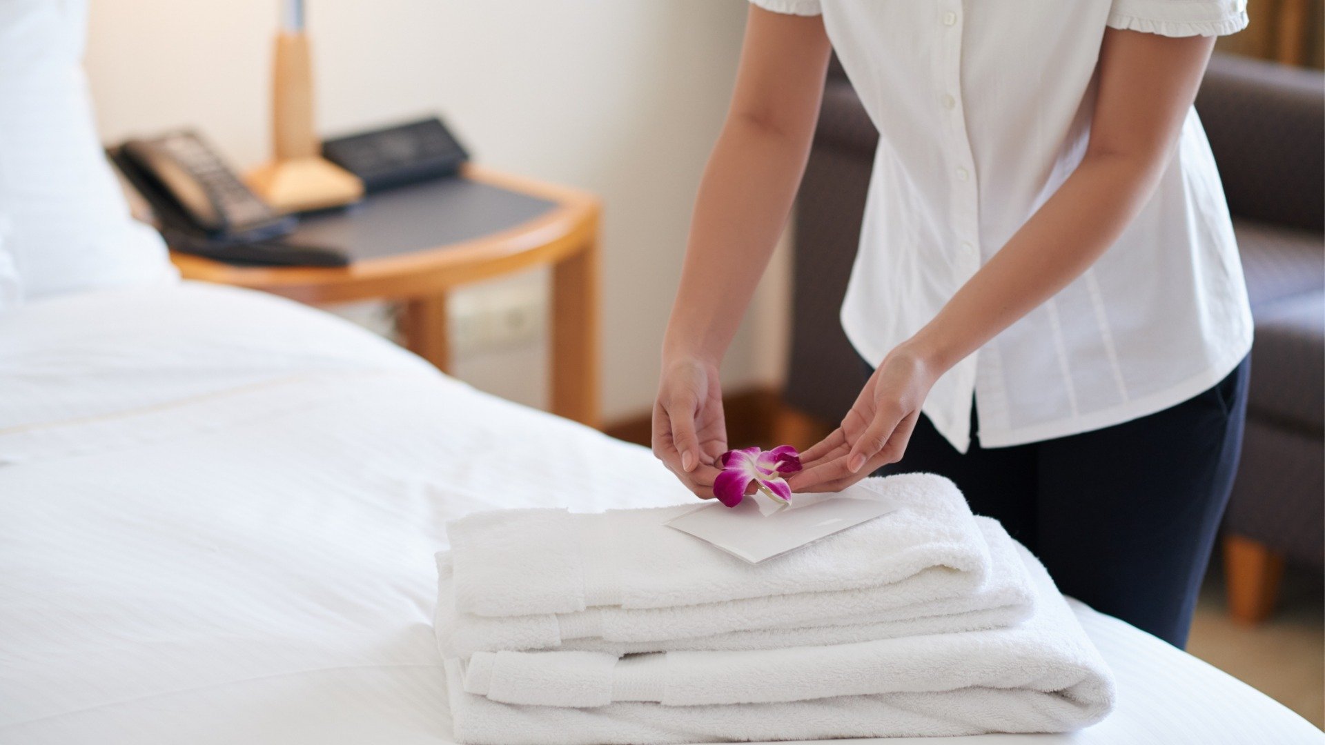 This image shows a woman placing a fresh flower and a note on a pile of towels on a hotel bed. 