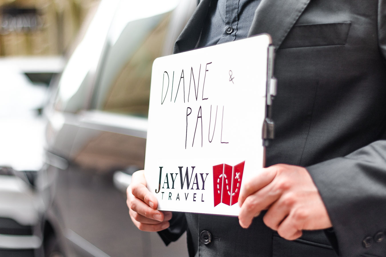 This is a close-up of a man standing outside a car and holding a sign that reads "Diane & Paul, JayWay Travel". 