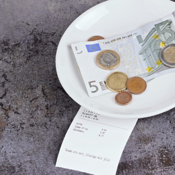 This is a top-down image that shows a small white plate with a 5 EUR bill and some euro coins on a grey table.
