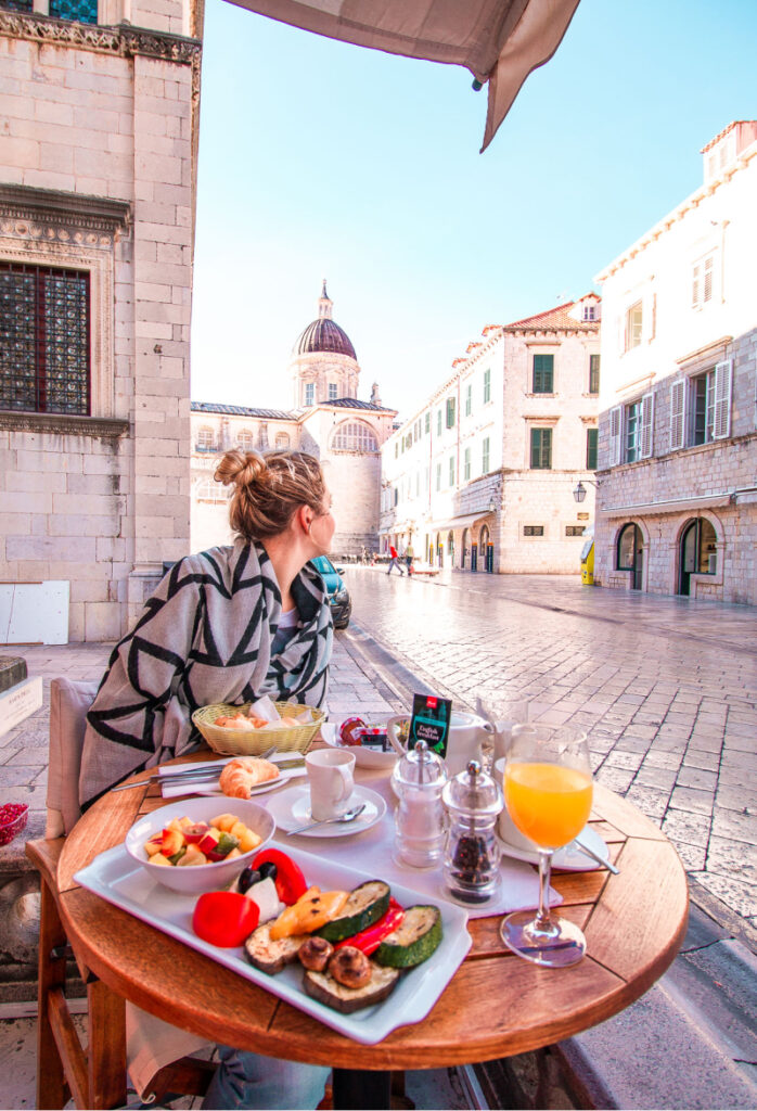 A woman sitting in a shawl enjoying an early breakfast in Dubrovnik's old town. On the table is orange juice, a plate of grilled vegetables and salt and pepper grinders.