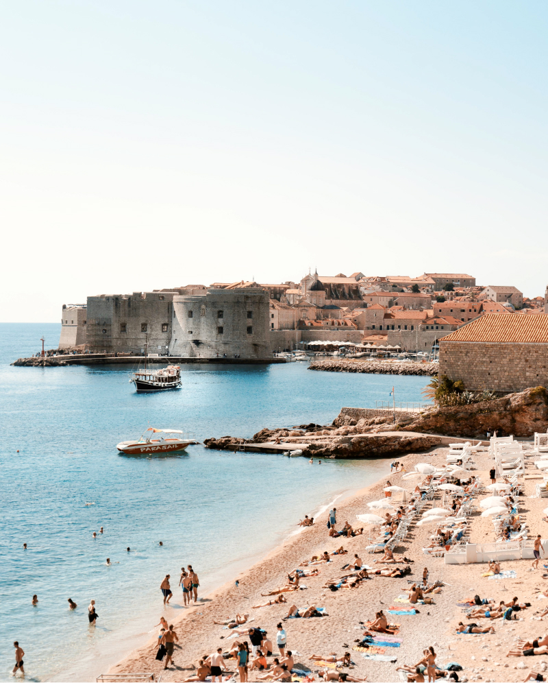 People sunbathing and paddling at Banje beach Dubrovnik, with the old city of Dubrovnik in the background, on a cloudless day.