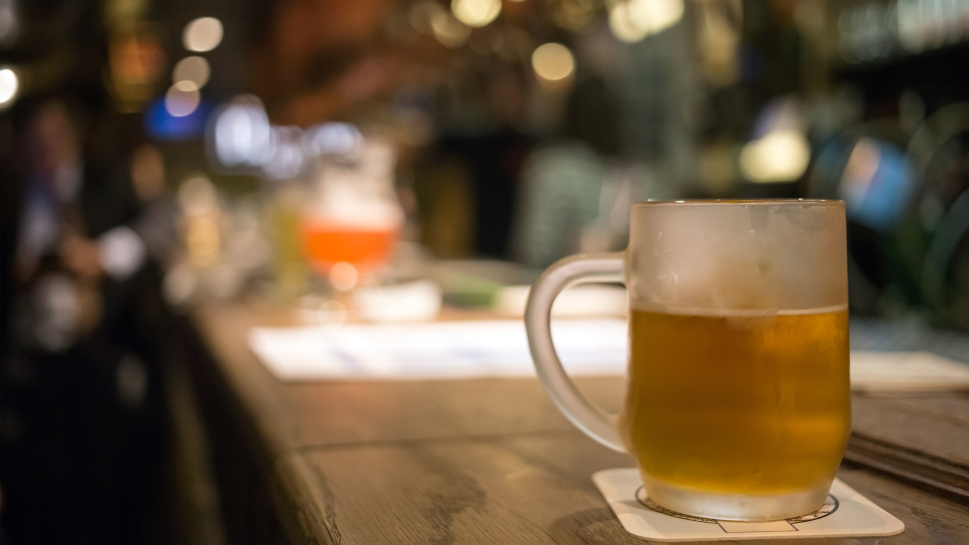 This image shows a frosty glass of beer on a bar counter with a blurry background. 