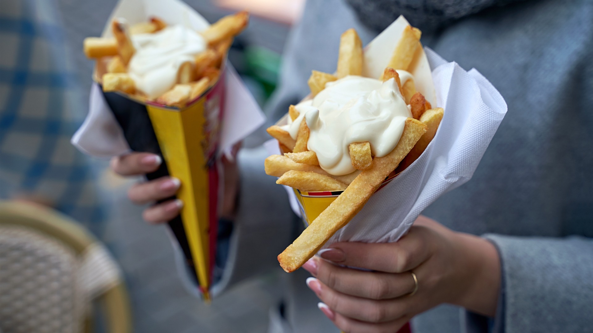 This image is a close-up of a woman holding two paper cones filled with Belgian frites, topped with mayonnaise sauce. Sampling Belgian frites is one of the main reasons to visit Brussels. 