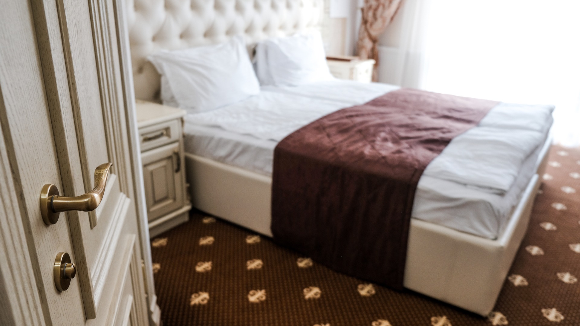 This image shows a hotel room with an old-fashioned bed and bedside table. The floor is covered by carpet from wall to wall. 