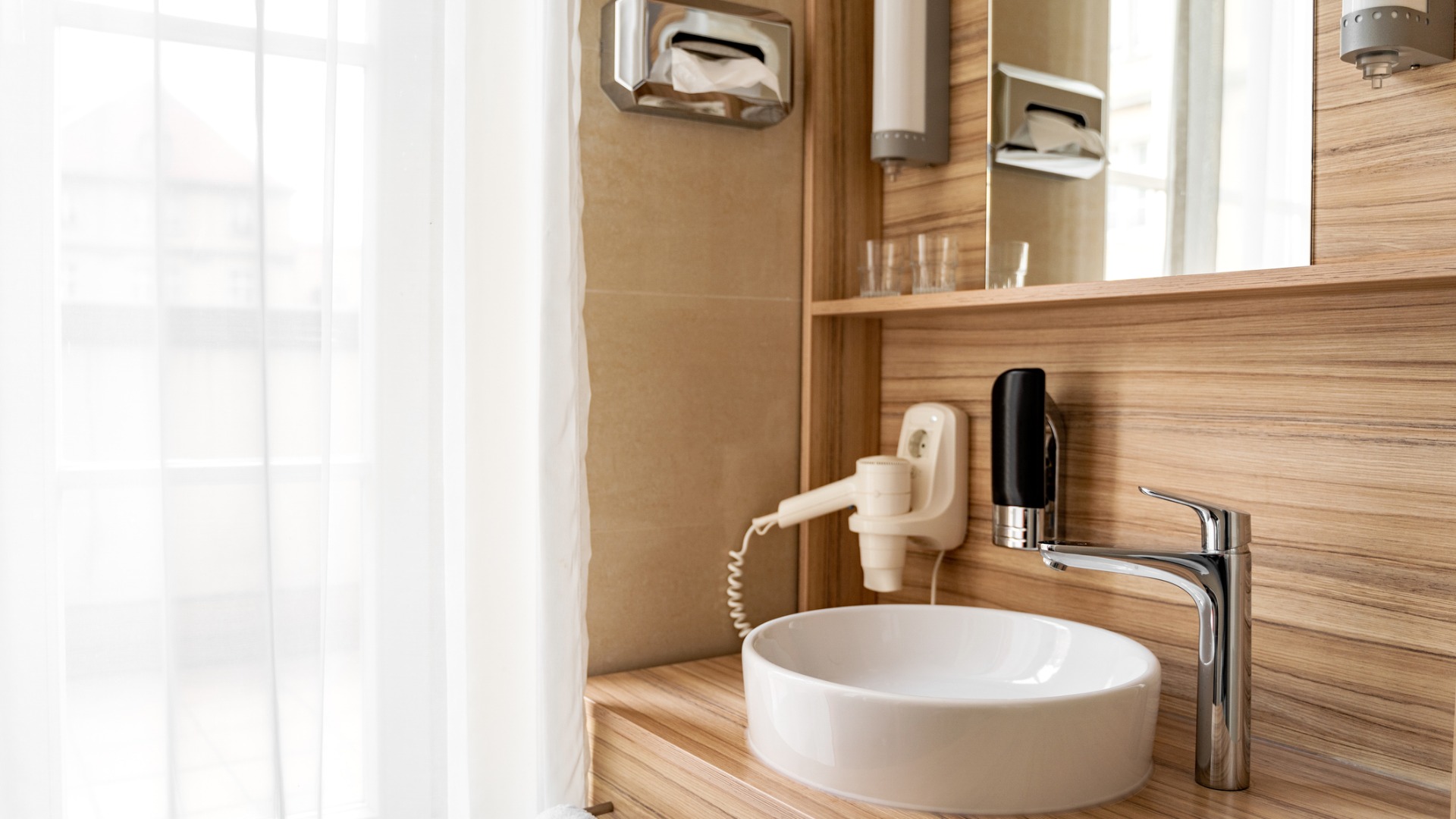 This image shows a small bathroom in a hotel room with window, sink, faucet, hair dryer and mirror.