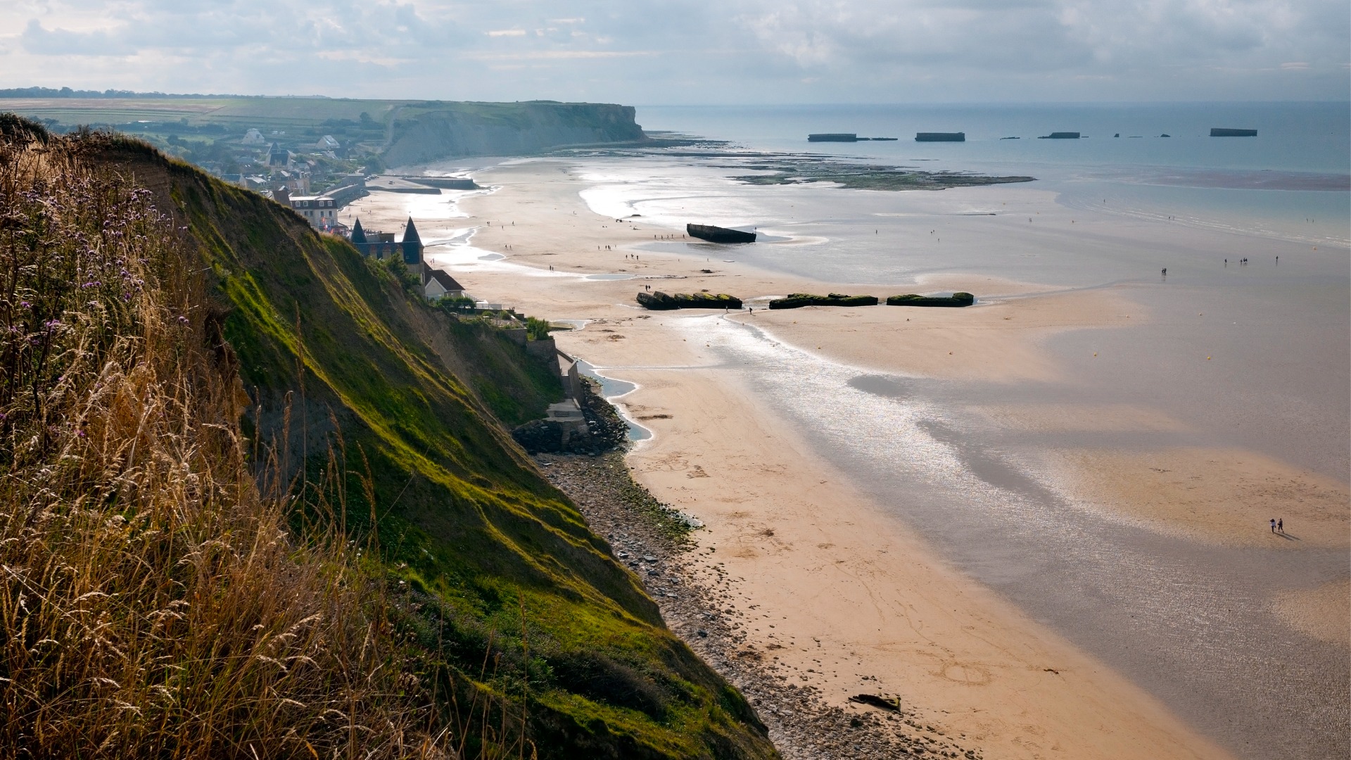This is a panoramic view of a beach in Normandy. There are green cliffs going all the way down to the white sand. 