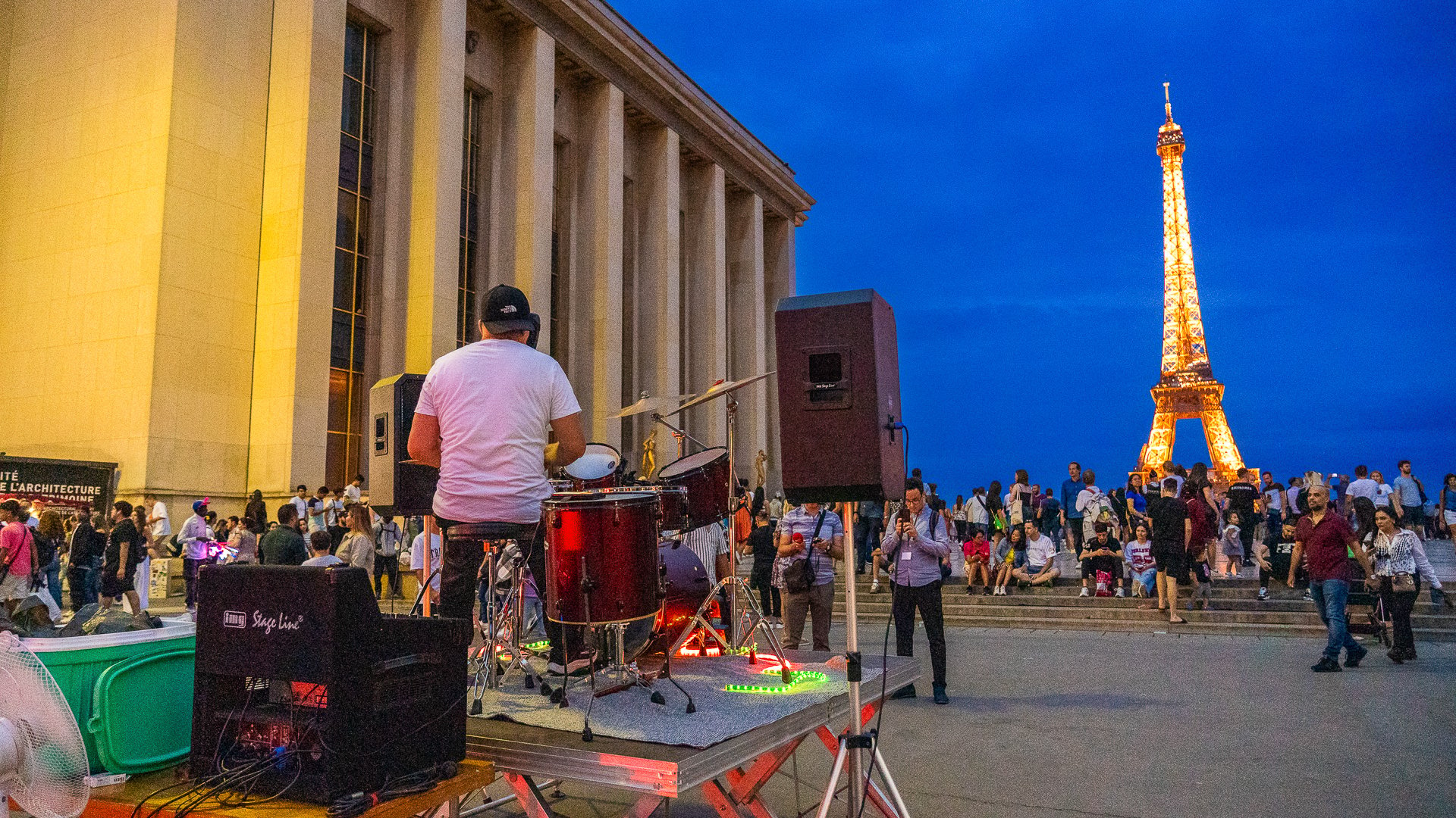 This image shows a band playing music with several people watching. The illuminated Eiffel Tower is in the background. 