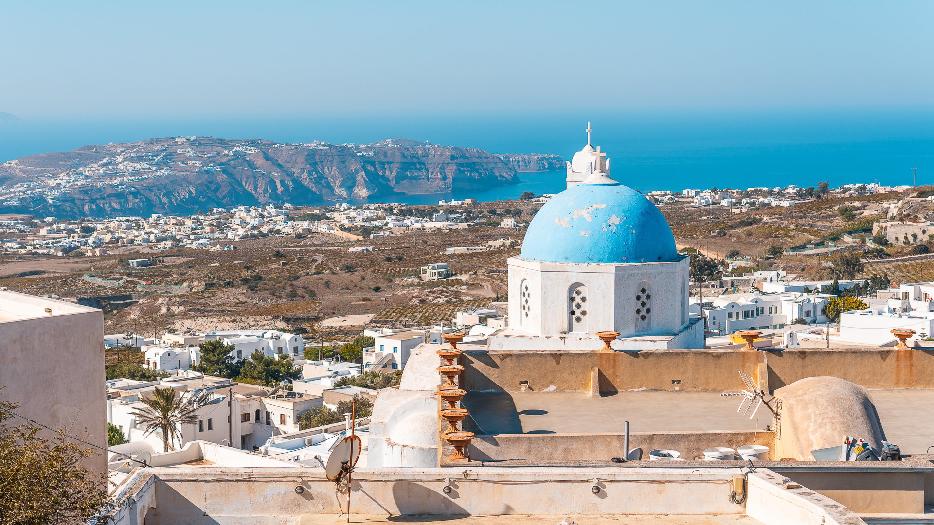 This is a panoramic view of Pyrgos village in Santorini with a blue-domed church in the foreground. 