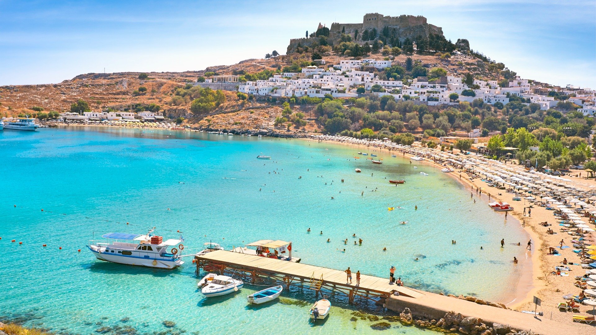 This is a panoramic view of Lindos. The Acropolis of Lindos is in the background and a beach with transparent waters takes up most of the photo. 