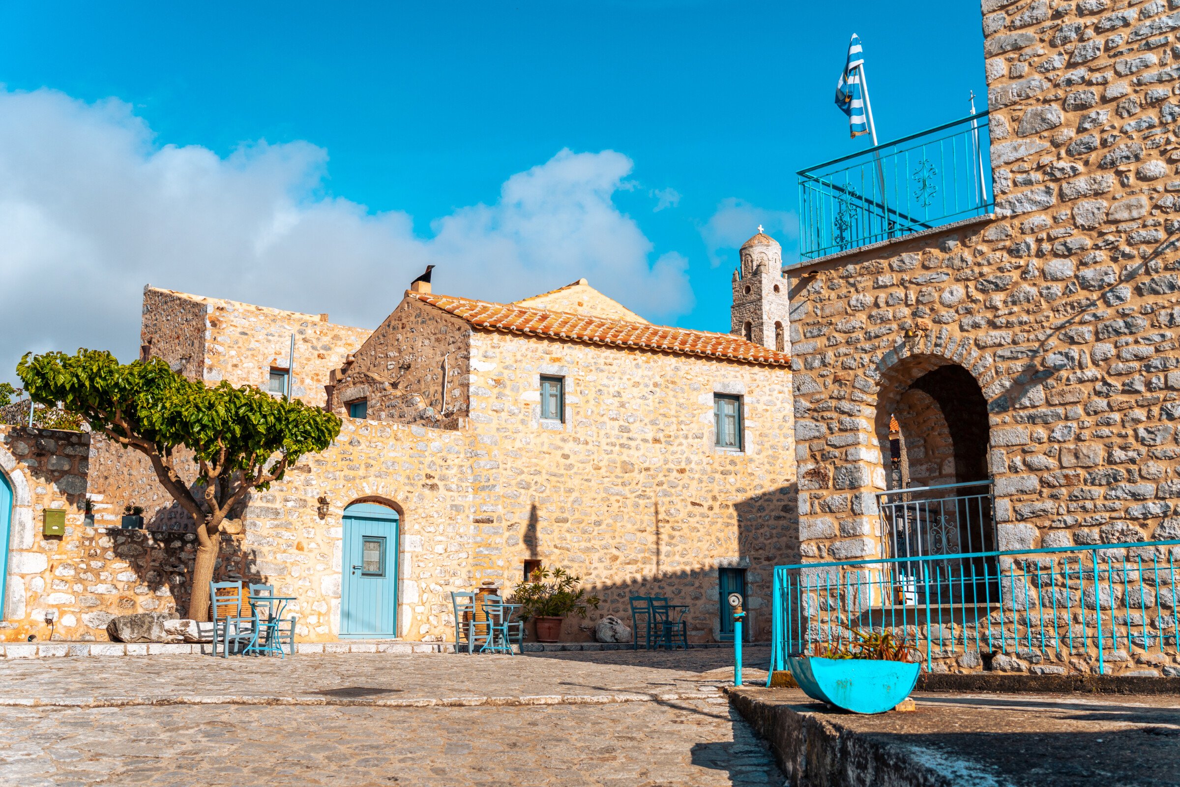 This image shows a peaceful square lined with stone buildings with sky blue shutters in Areopoli. 