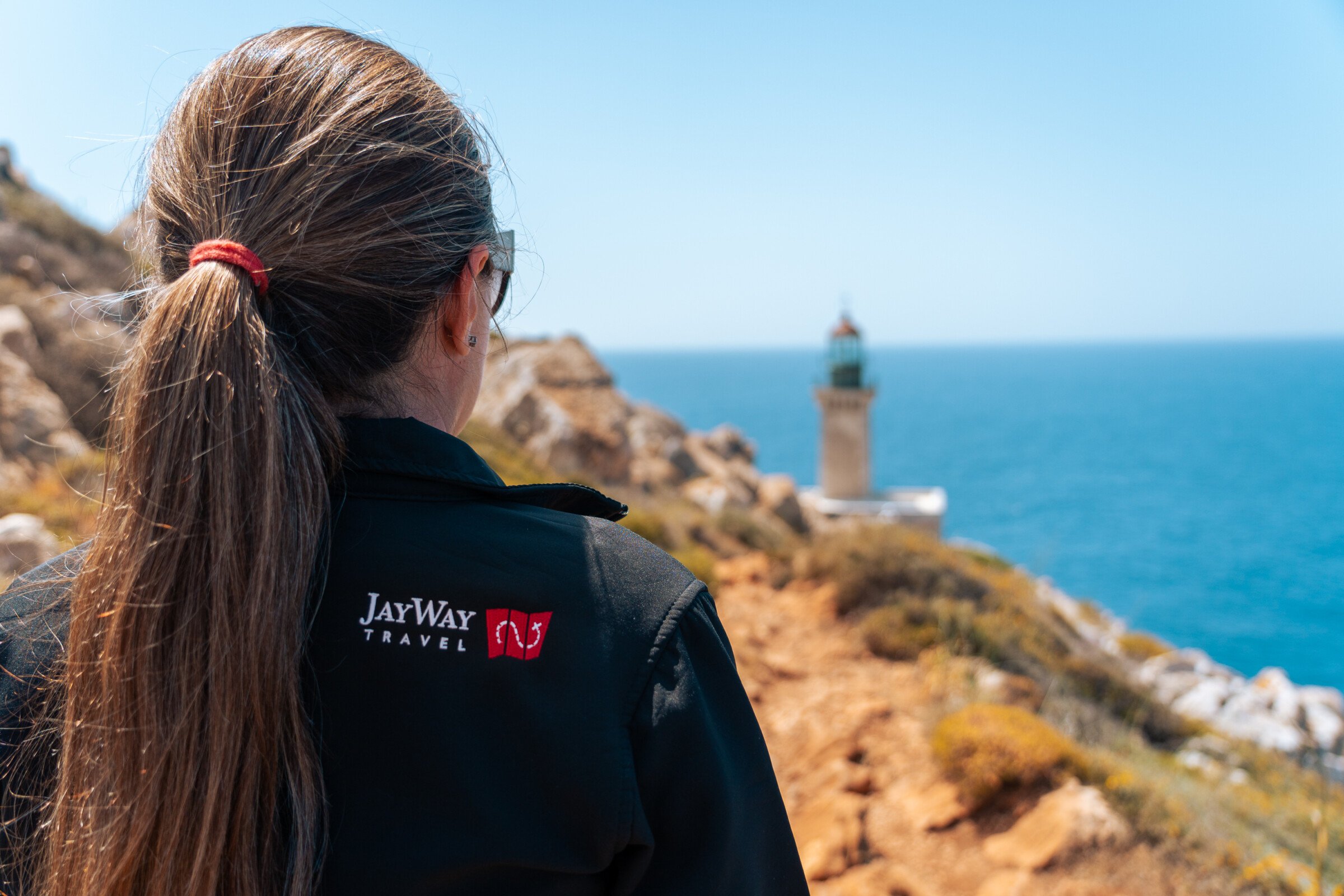 This image shows a woman with her back turned to the camera looking at the lighthouse of Cape Tainaro in the background. She's wearing a jacket with the JayWay Travel logo. 