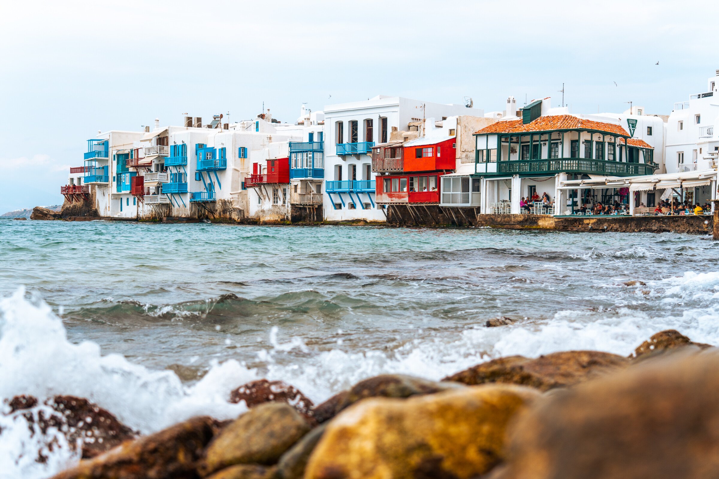 This image shows Little Venice in Mykonos with its characterisitic white houses with colorful wooden balconies built on the edge of the sea. 
