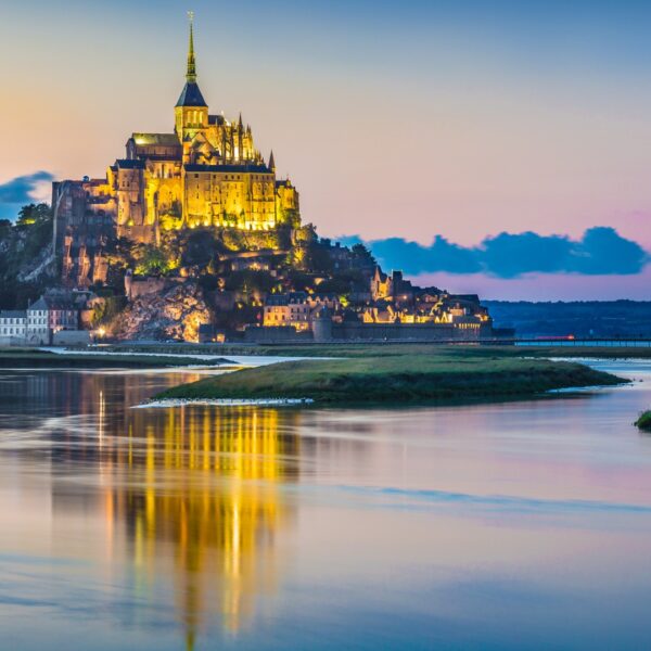 This image shows the tidal island of Mont-Saint-Michel dimly lit at twilight. The tiny rocky island is donimated by the enormous and impressive abbey that sits atop it.