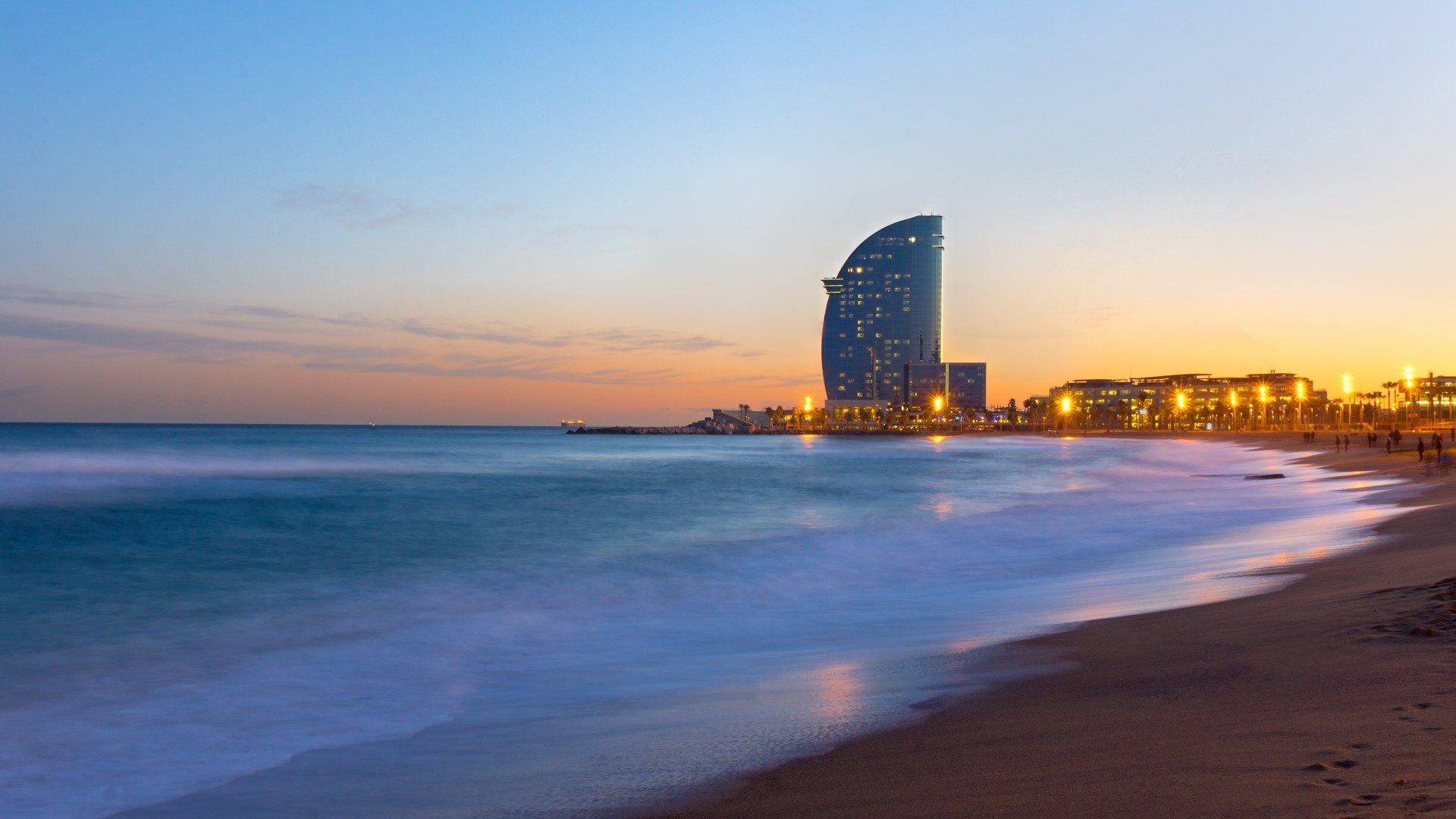In the background, this image shows the iconic sail-shaped building that dominates La Barceloneta. In the foreground, the waves and the sand dyed in sunset colors. 