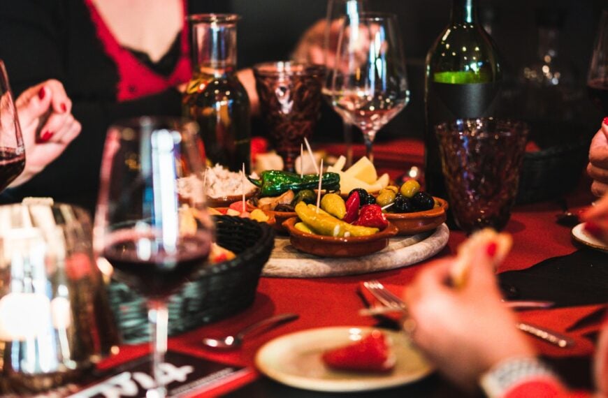 This image shows a table set with a large tray of tapas in the middle, a bottle and glasses of wine, and the hands of people around the table eating.