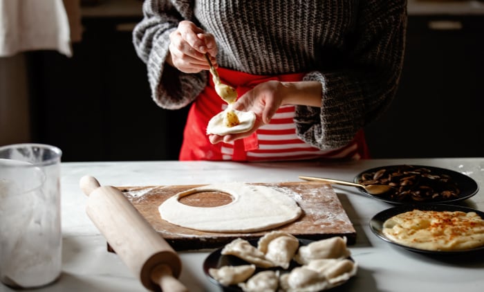 A person wearing a gray sweater and red apron is preparing stuffed dumplings in a kitchen They are spooning filling onto a circular piece of dough placed on a floured wooden board Various ingredients and finished dumplings are arranged nearby along with a rolling pin and a plate of sauted mushrooms