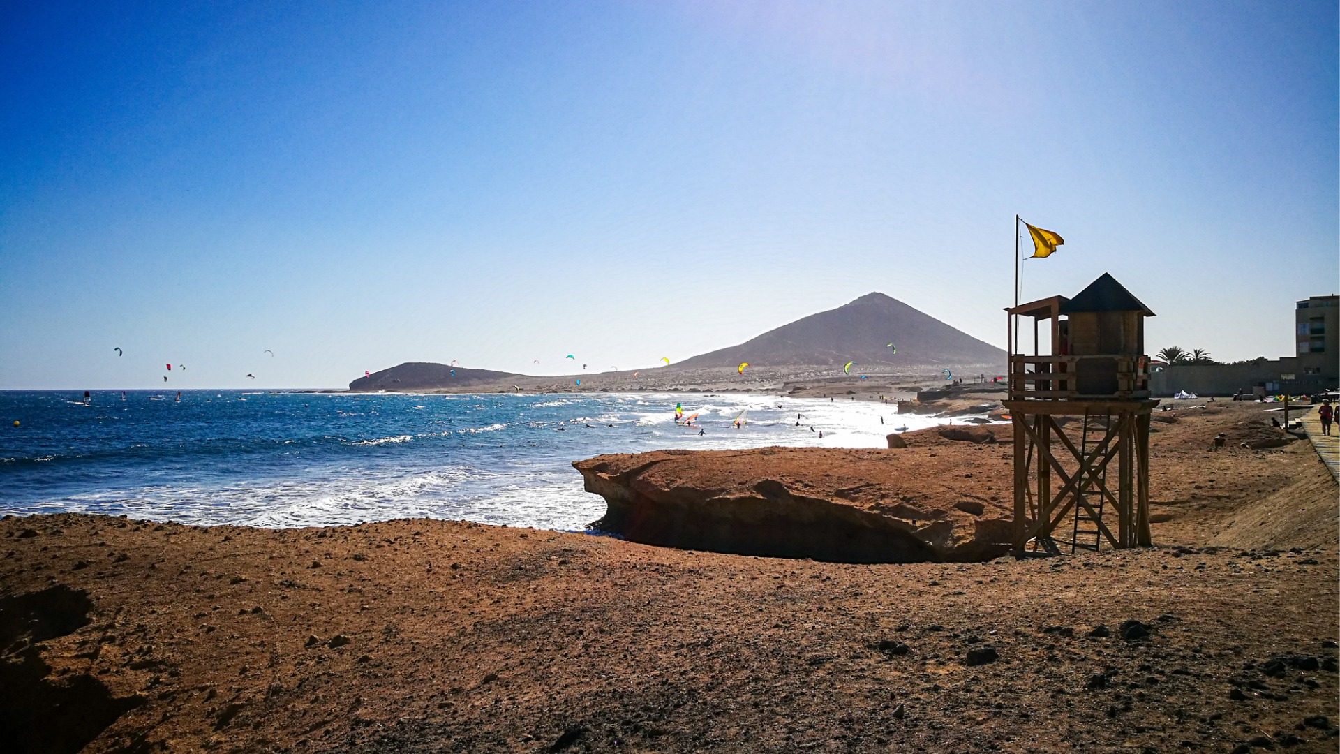 A red-sanded beach with many kite surfers in the background.