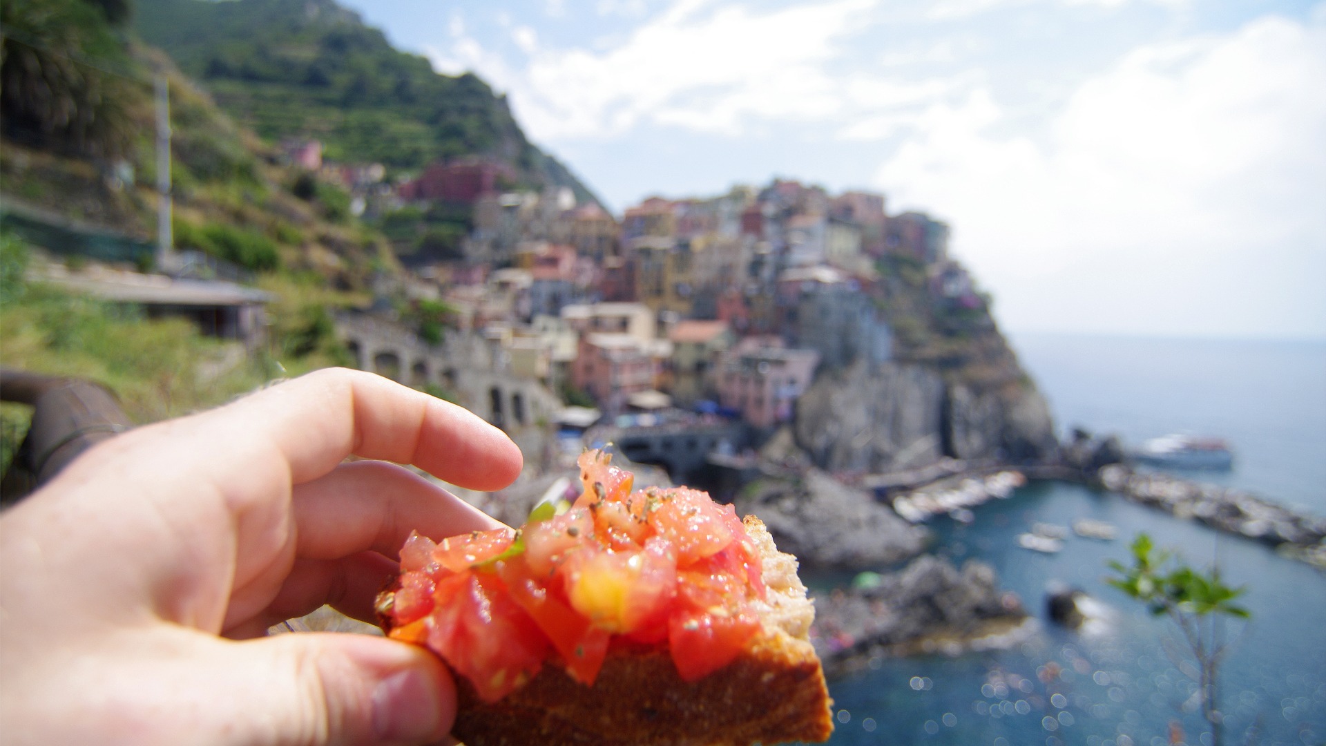 A hand holding a bruschetta in the foreground with a typically Italian landscape in the background.
