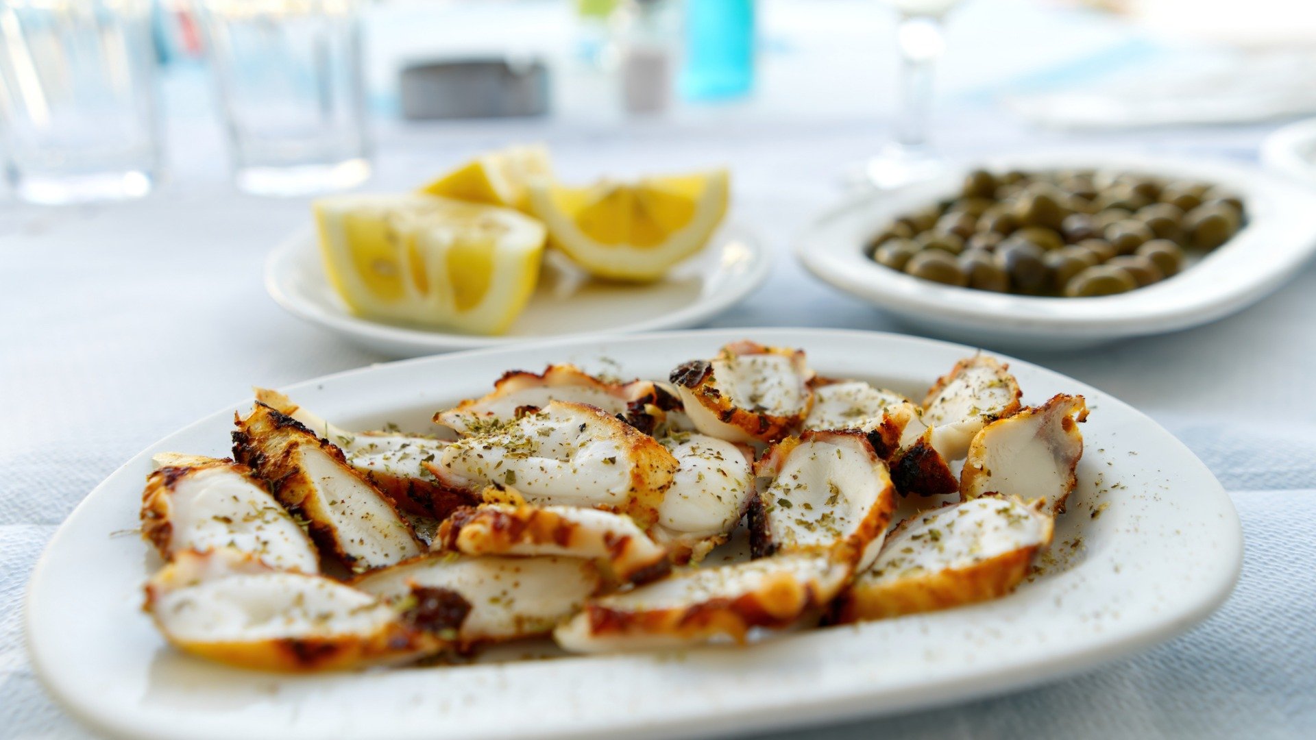 In the foreground, a white plate with pieces of grilled octopus. In the background, a small plate with lemon wedges and another with olives. 