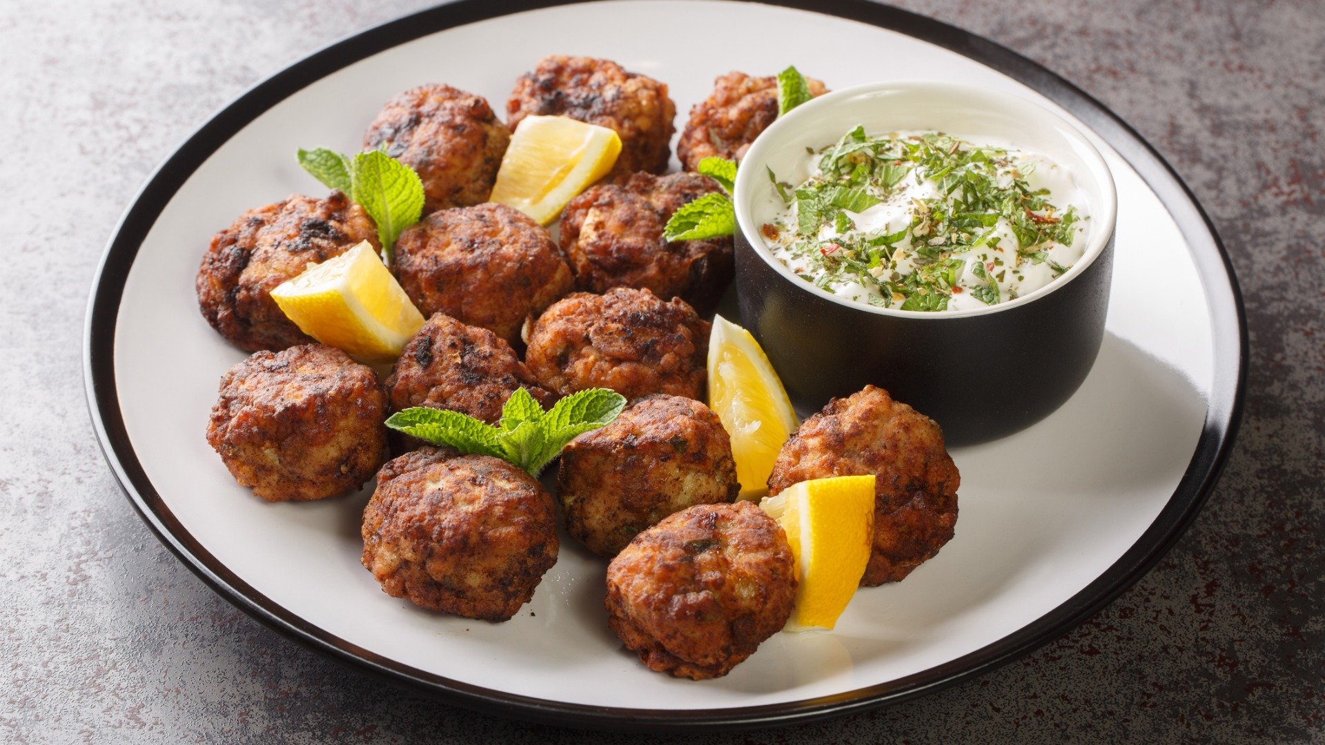 A plate with many meatballs and a smaller bowl of Tzatziki dip on it. 