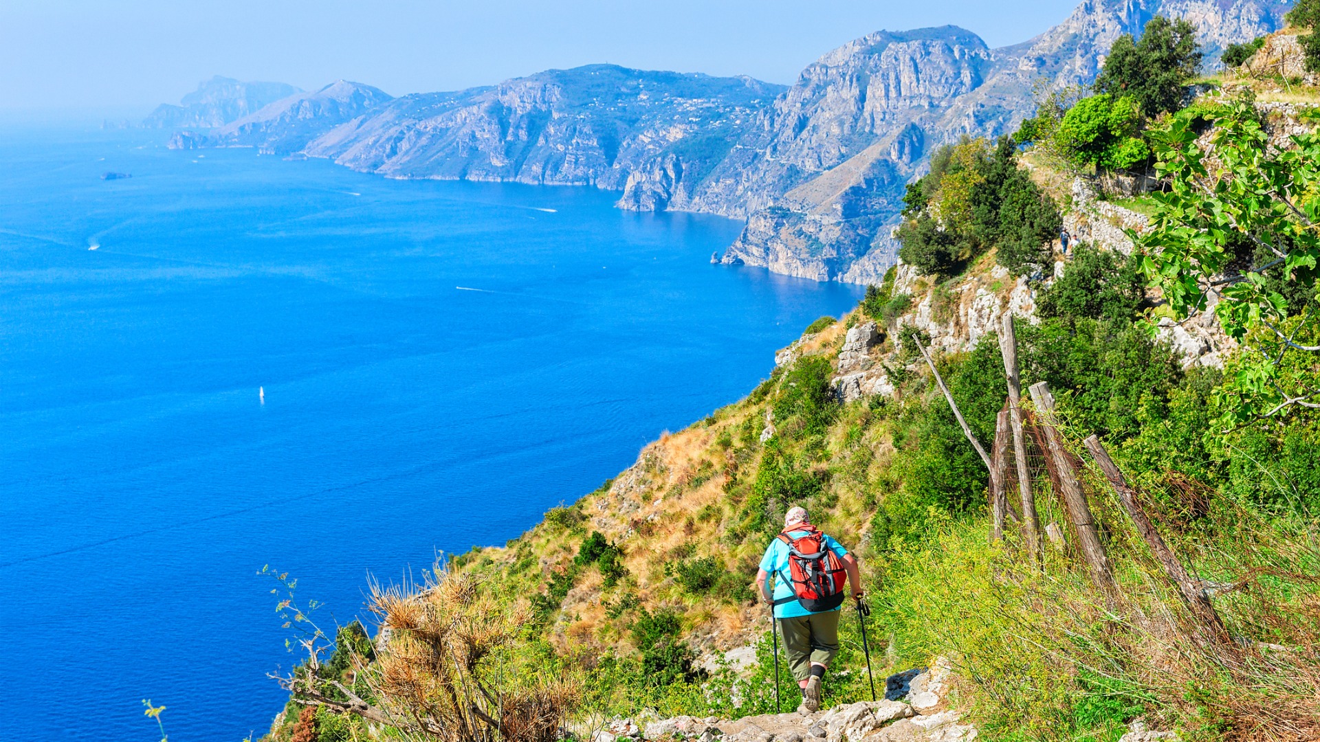 A man with a backpack is hiking along the Path of Gods. Great sea views and tall mountains in the background. 