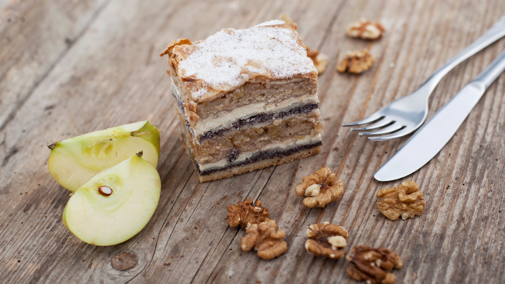 A piece of Prekmurska Gibanica, two apple slices, walnuts, a knife, and a fork on a wooden surface. 