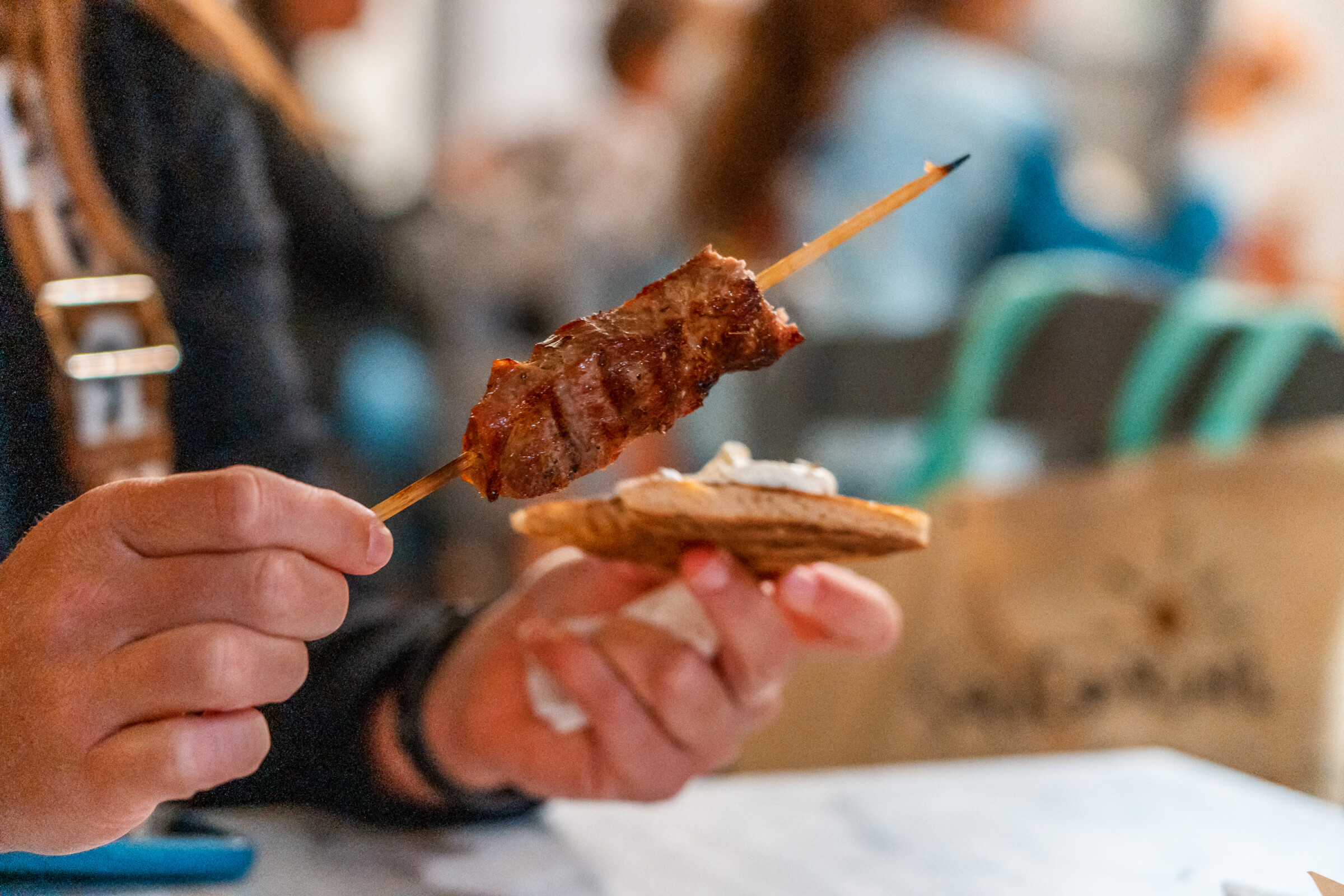 Two hands, one is holding a Souvlaki skewer, the other a small piece of Pita bread. 