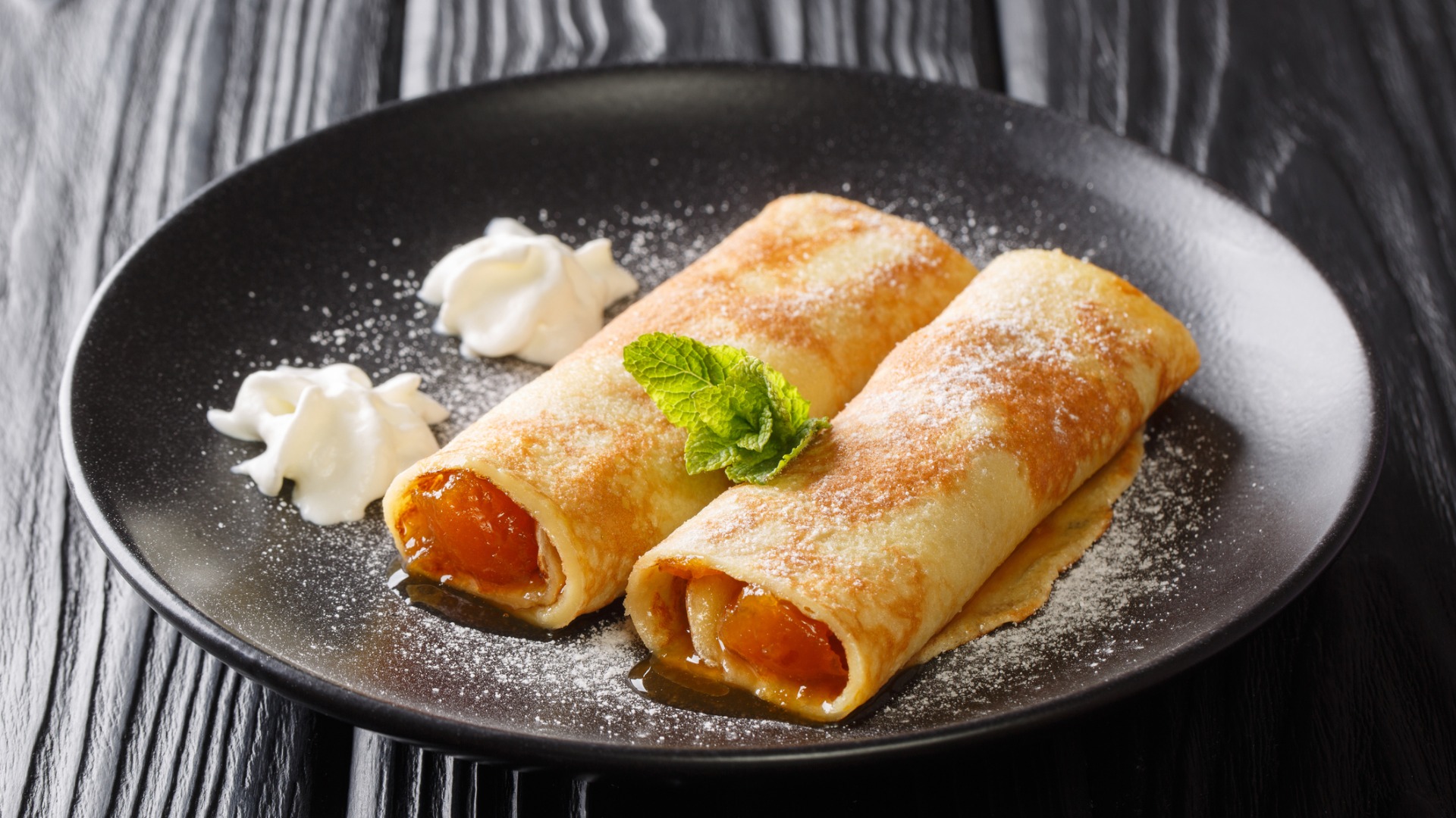 A close-up of a black plate with two Viennese crepes filled with apricot jam and served with whipped cream on the side. 