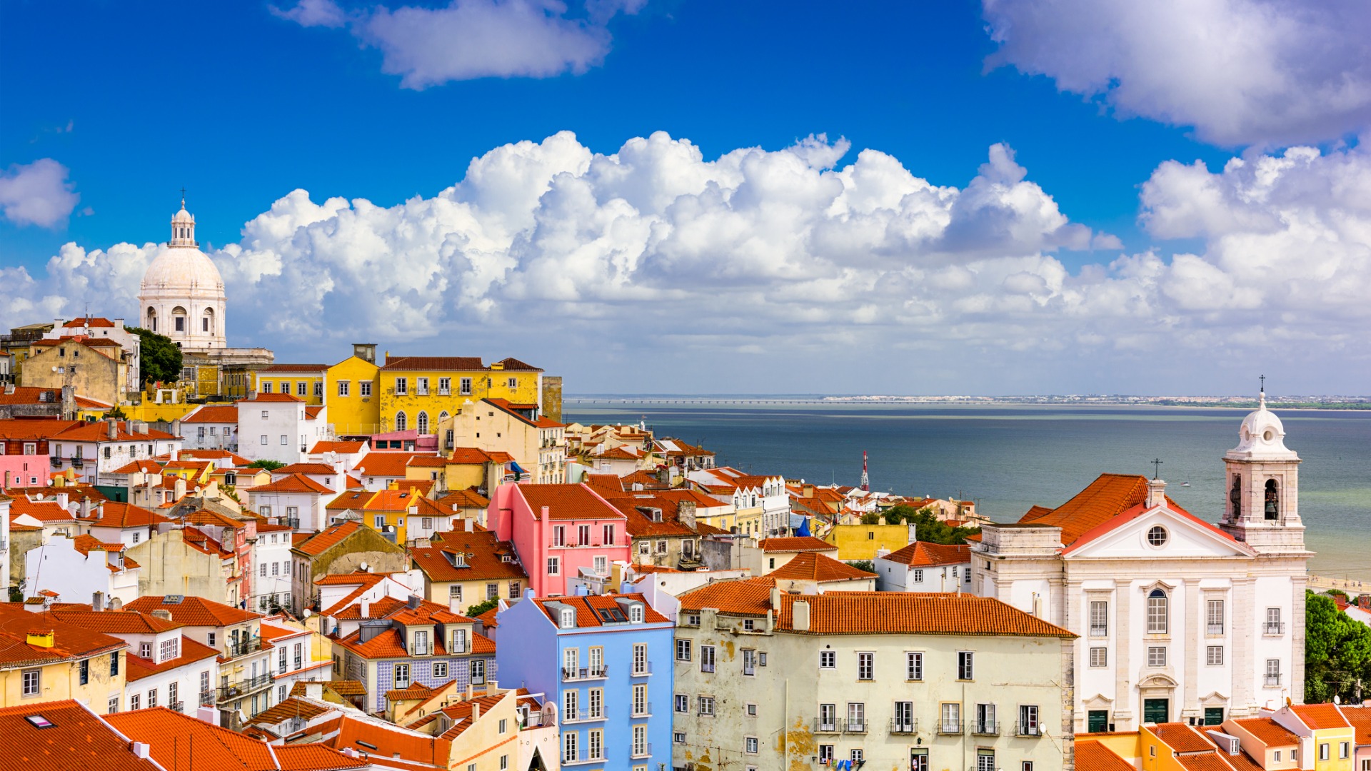 A view of Lisbon's red rooftops with the Tagus River in the background under a cloudy yet bright sky. 