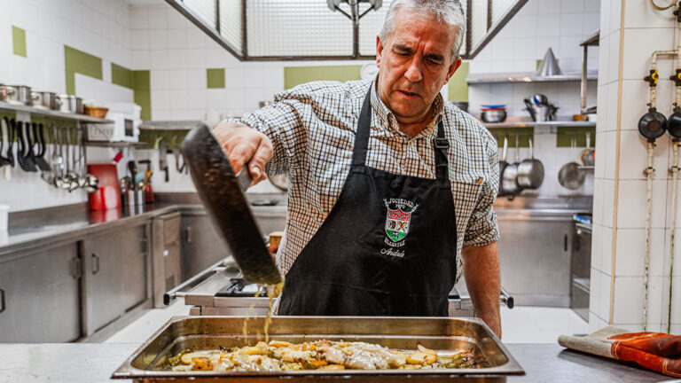 An older man in a checkered shirt and an apron is cooking in a professional kitchen He is carefully pouring liquid from a pan over a tray of food possibly preparing a baked dish